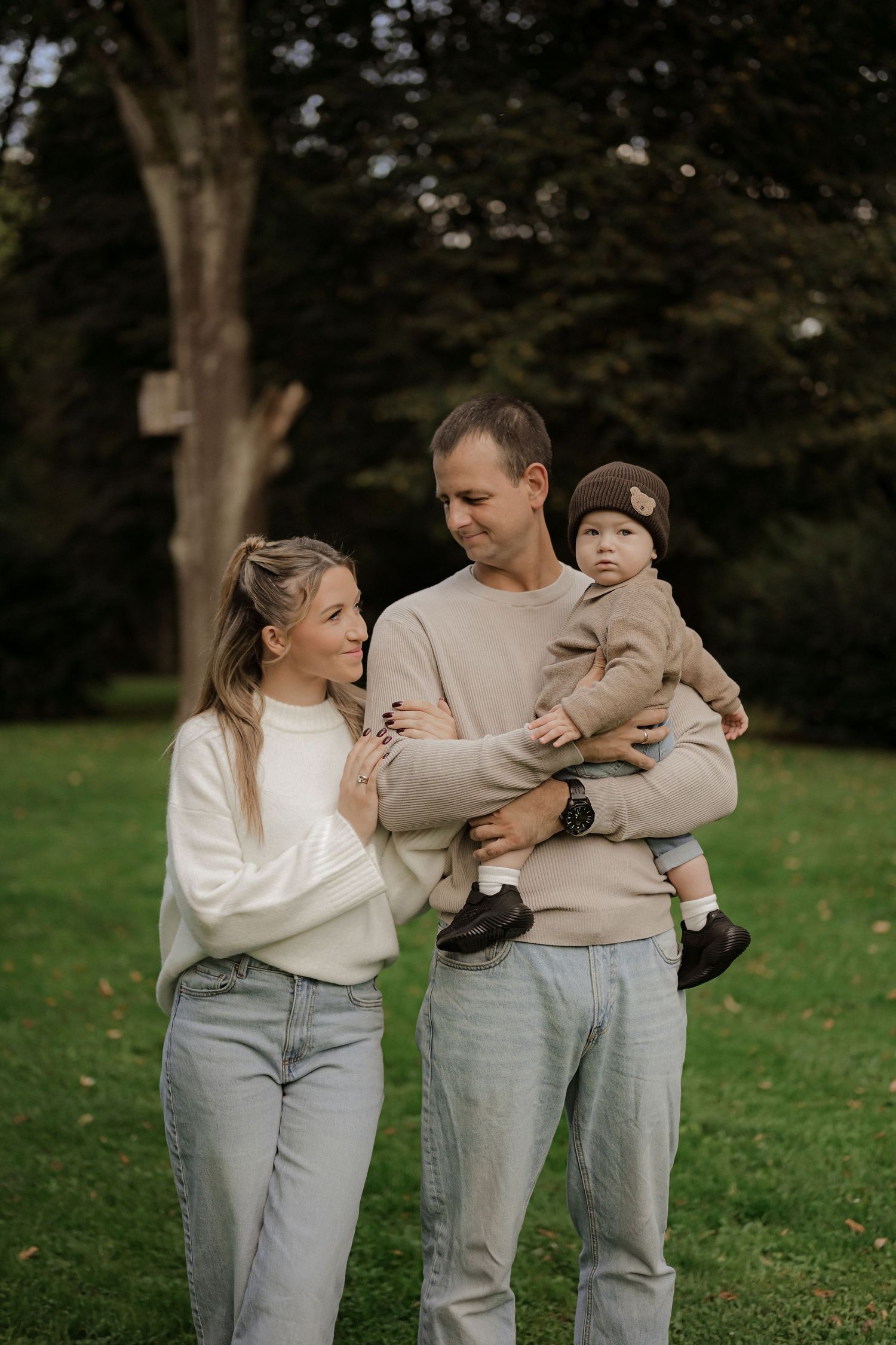 A young family with a baby poses outdoors on green grass with trees in the background.