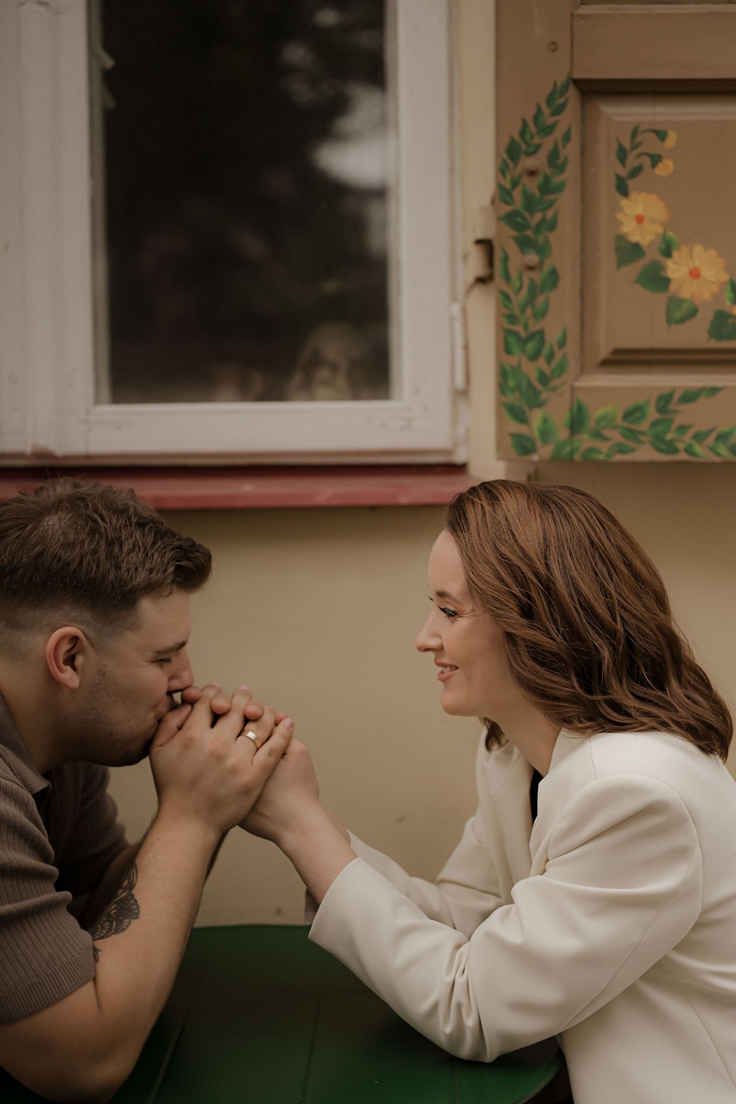 A romantic couple sits at a table as a man gently kisses a smiling woman's hand, with a floral painted cabinet behind.