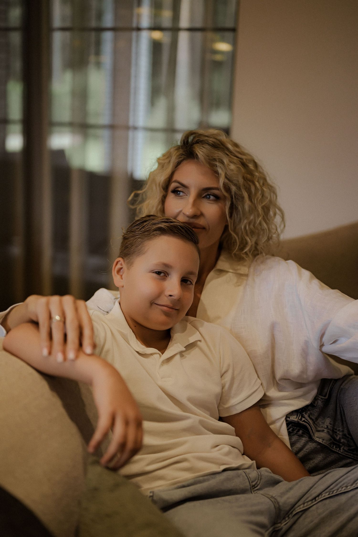 A curly-haired blonde woman and young boy smile together on a couch in a warm, cozy living room.