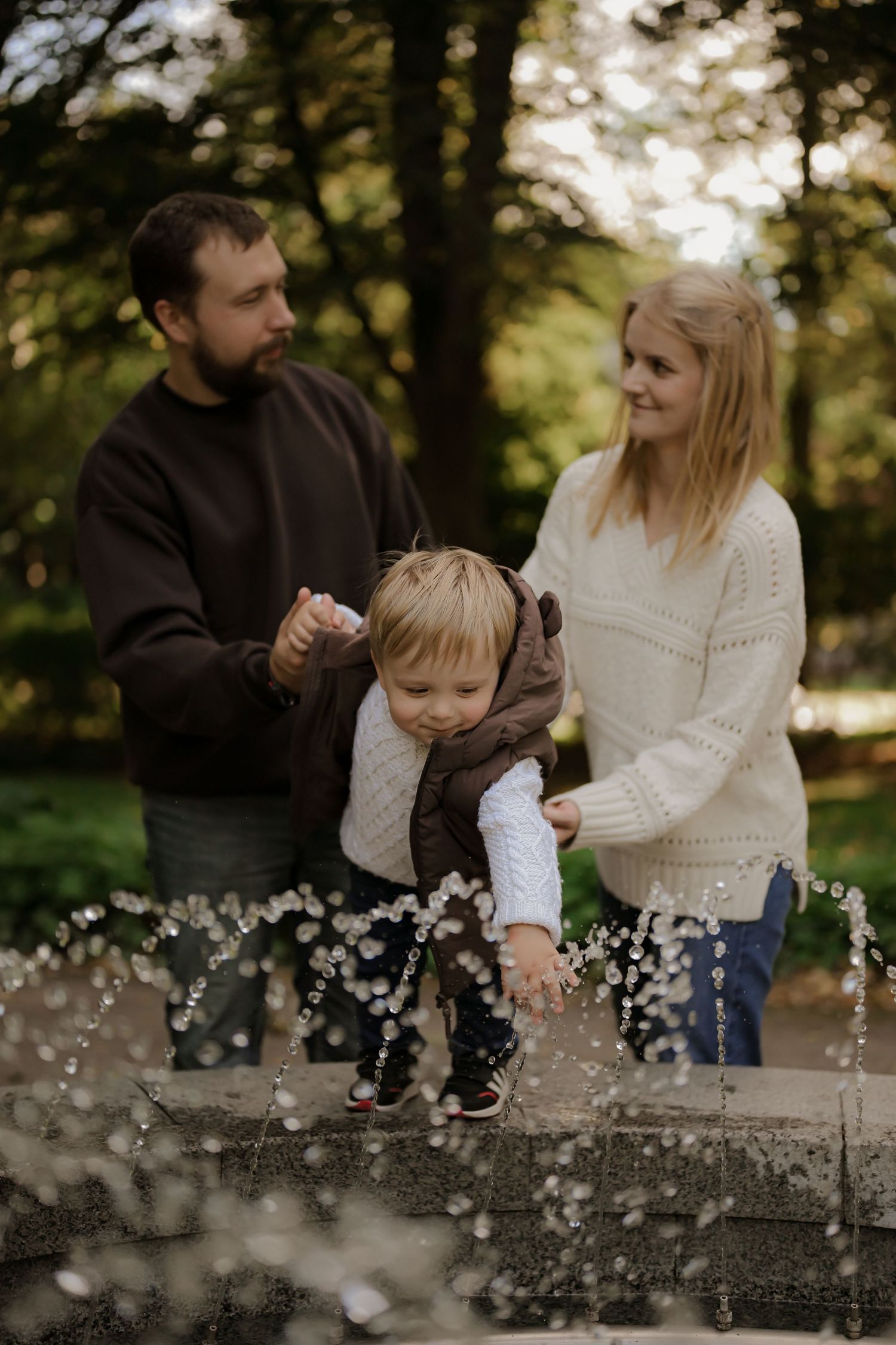 A young family with a toddler enjoys a sunny day by a fountain in a lush green park.