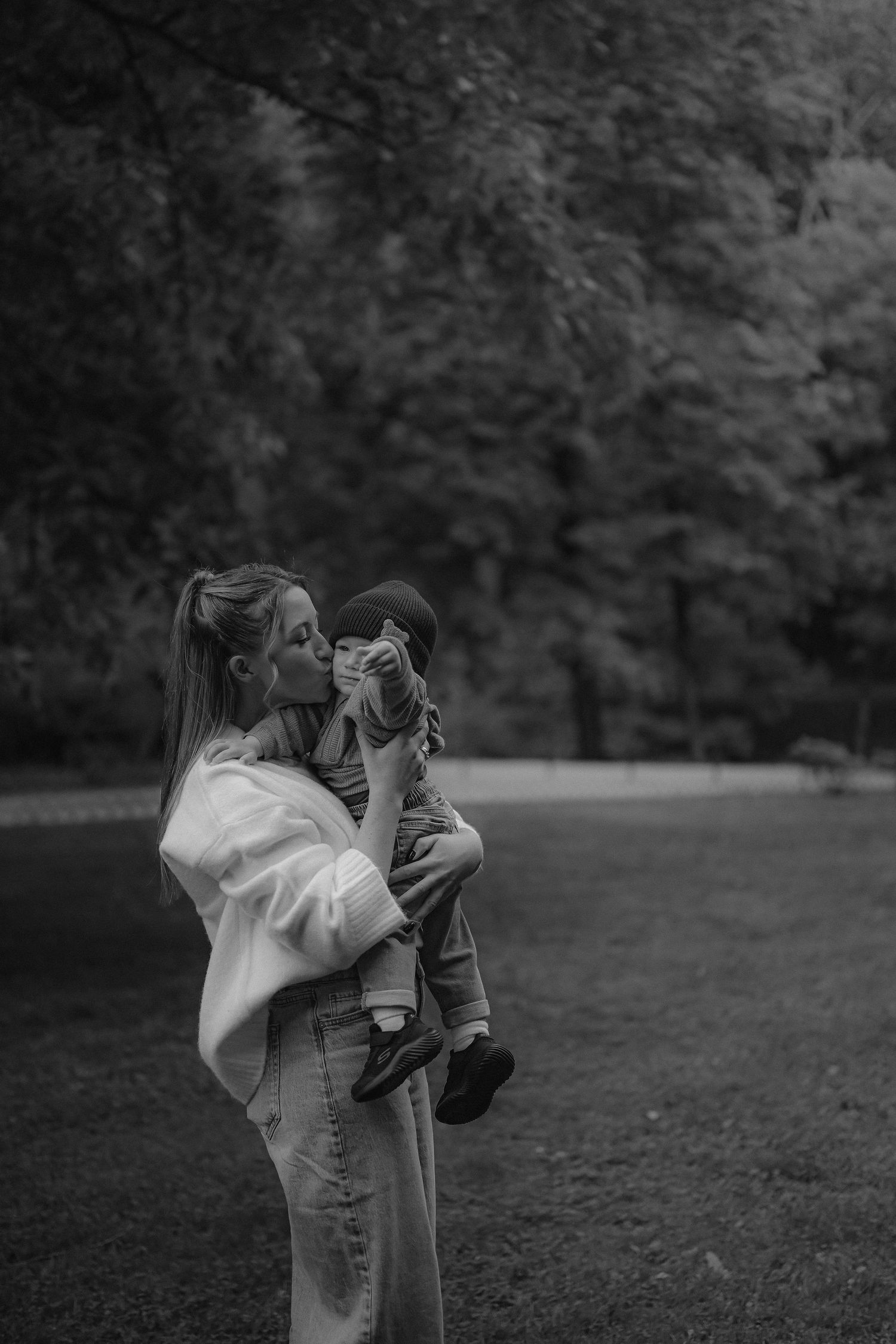 Black and white photo of a mother kissing her young child while holding them in a lush green park.