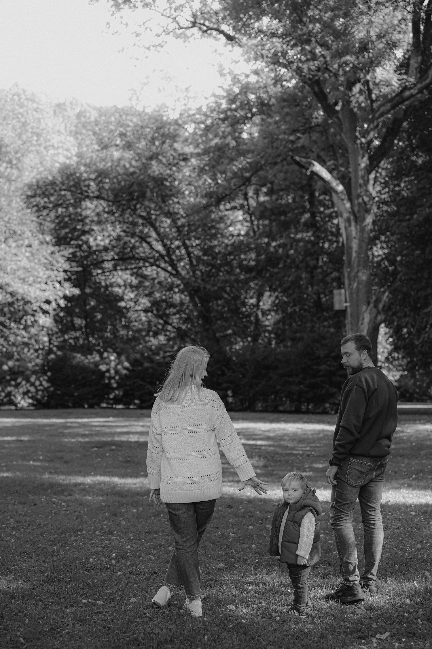 Black and white photo of a family with a young child walking together in a lush green park on a sunny day.