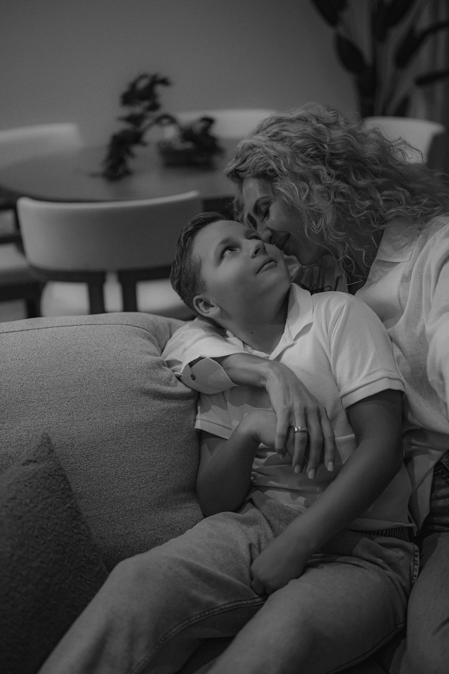 Black and white photo of a mother with curly hair kissing her young son on a cozy sofa at home.