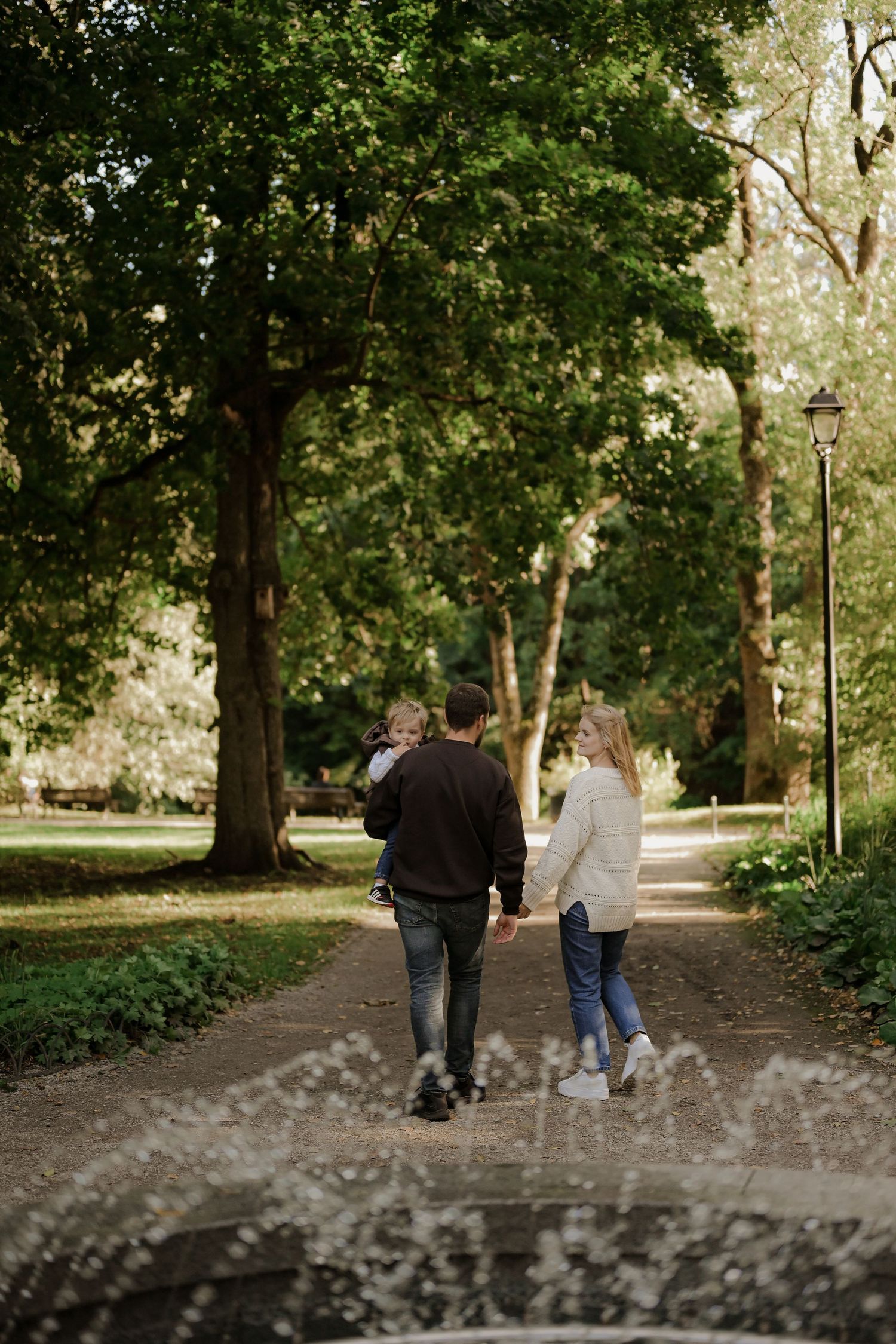 Young family walking hand in hand down a tree-lined park path, father carrying toddler.