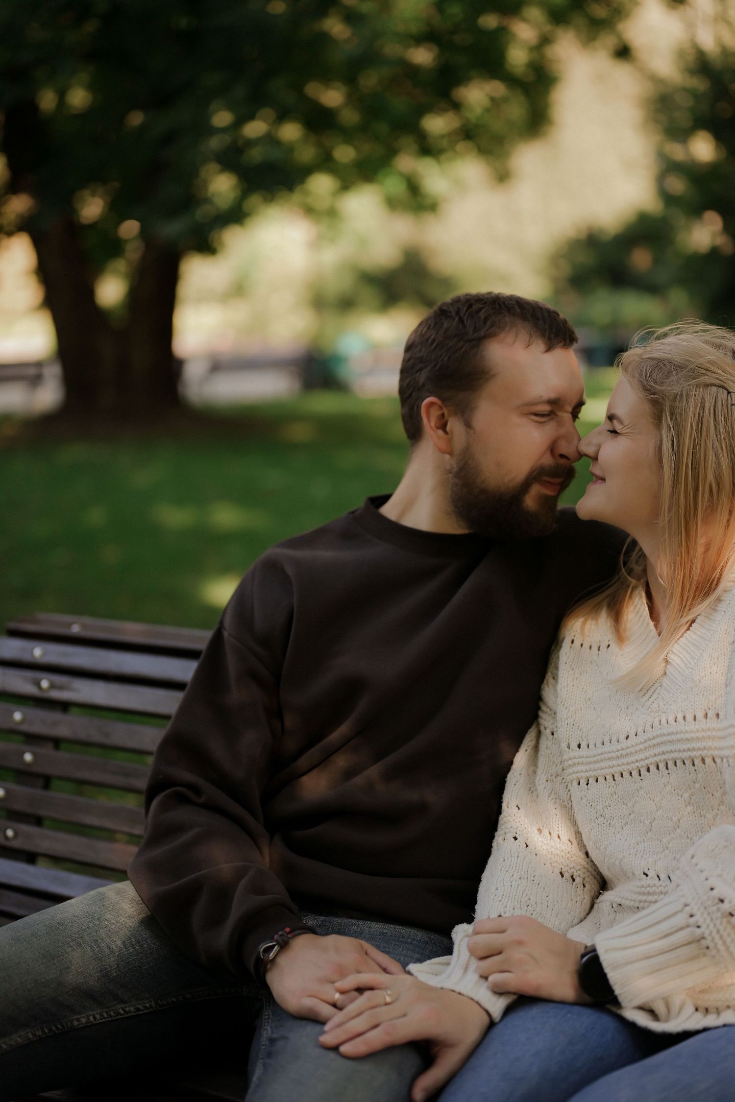 Couple sitting close on a park bench, sharing a romantic moment outdoors with lush green trees in the background.