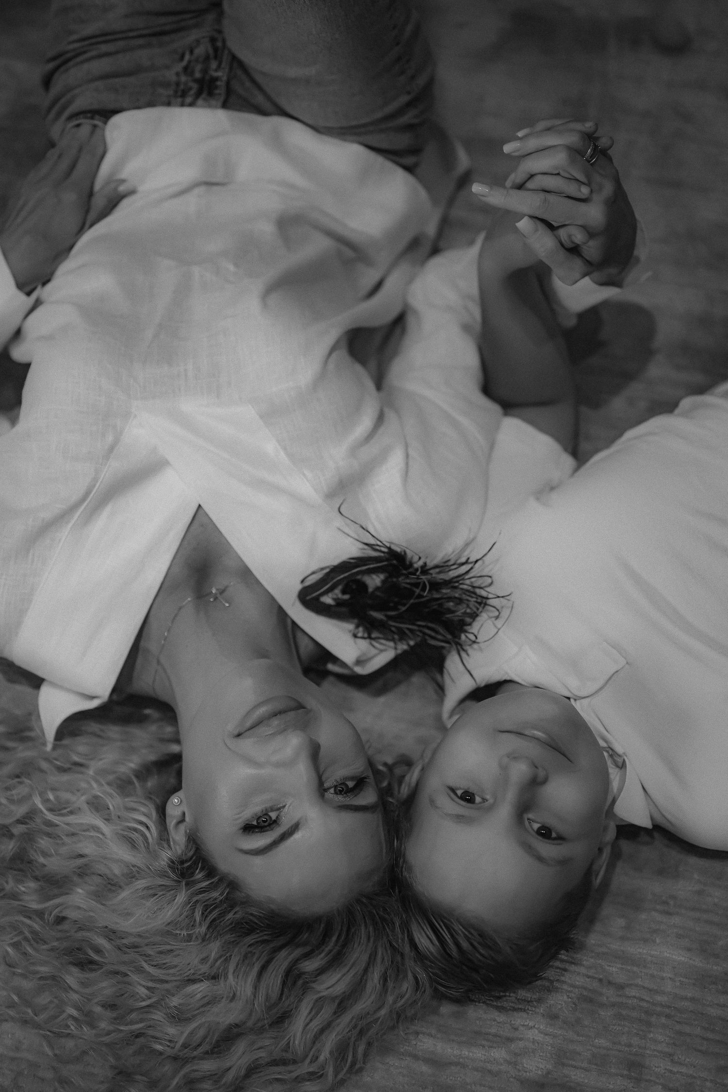 Black and white photo of two women lying on the floor, heads together, wearing white dresses, holding hands.