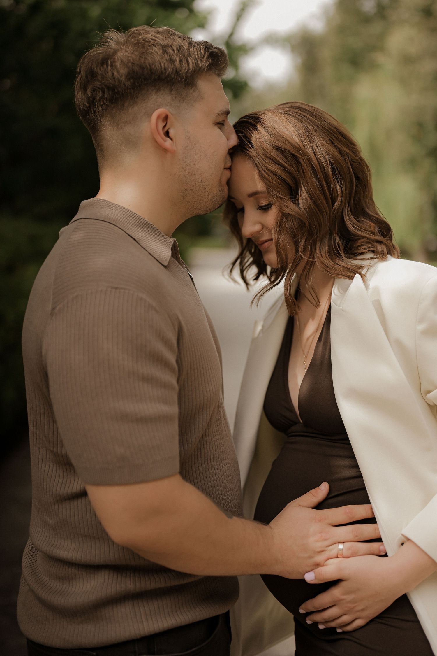 Couple sharing a tender moment, man kissing pregnant woman's forehead while cradling her baby bump outdoors.