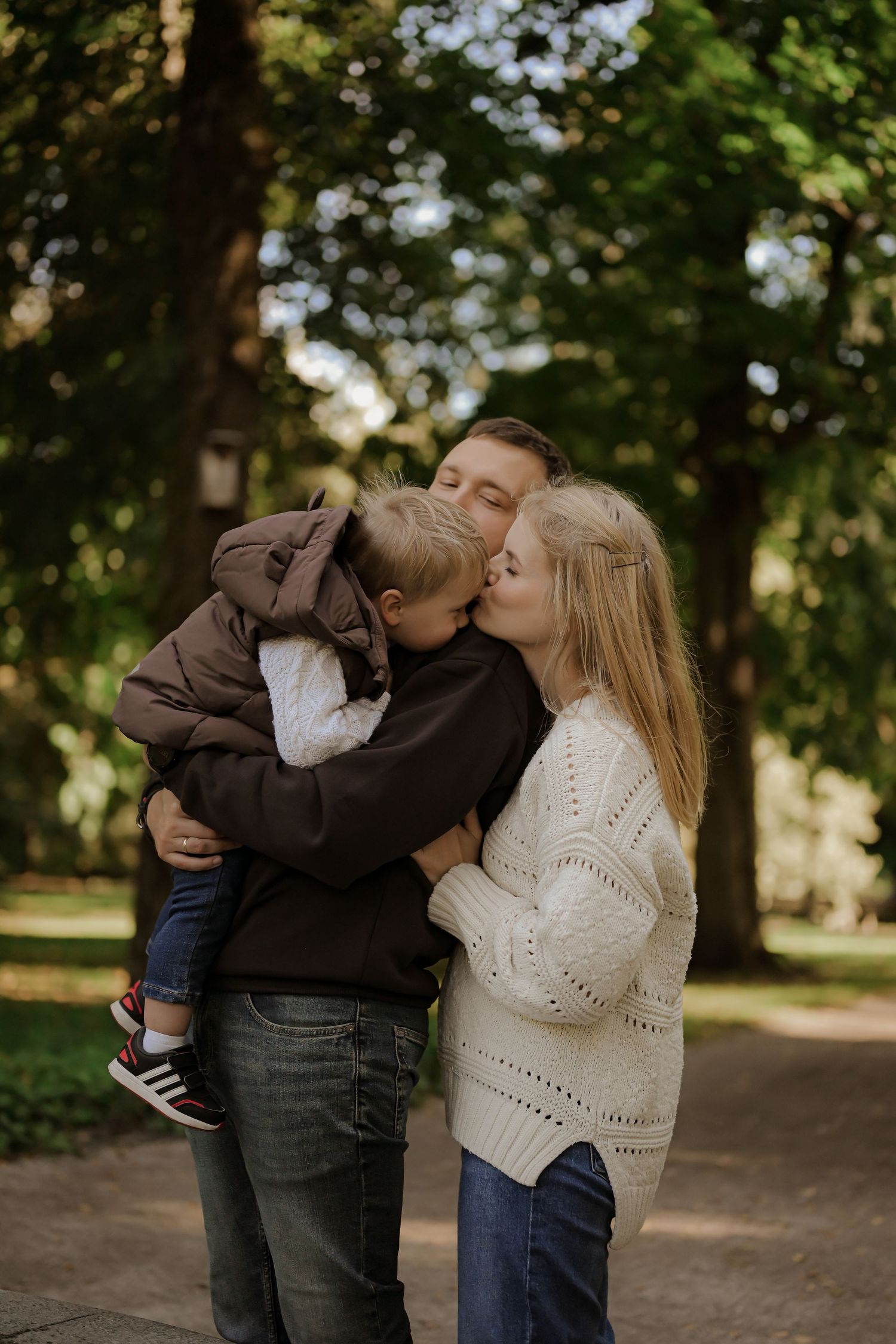 A loving family of three sharing kisses in a green park, with parents kissing their young child outdoors.