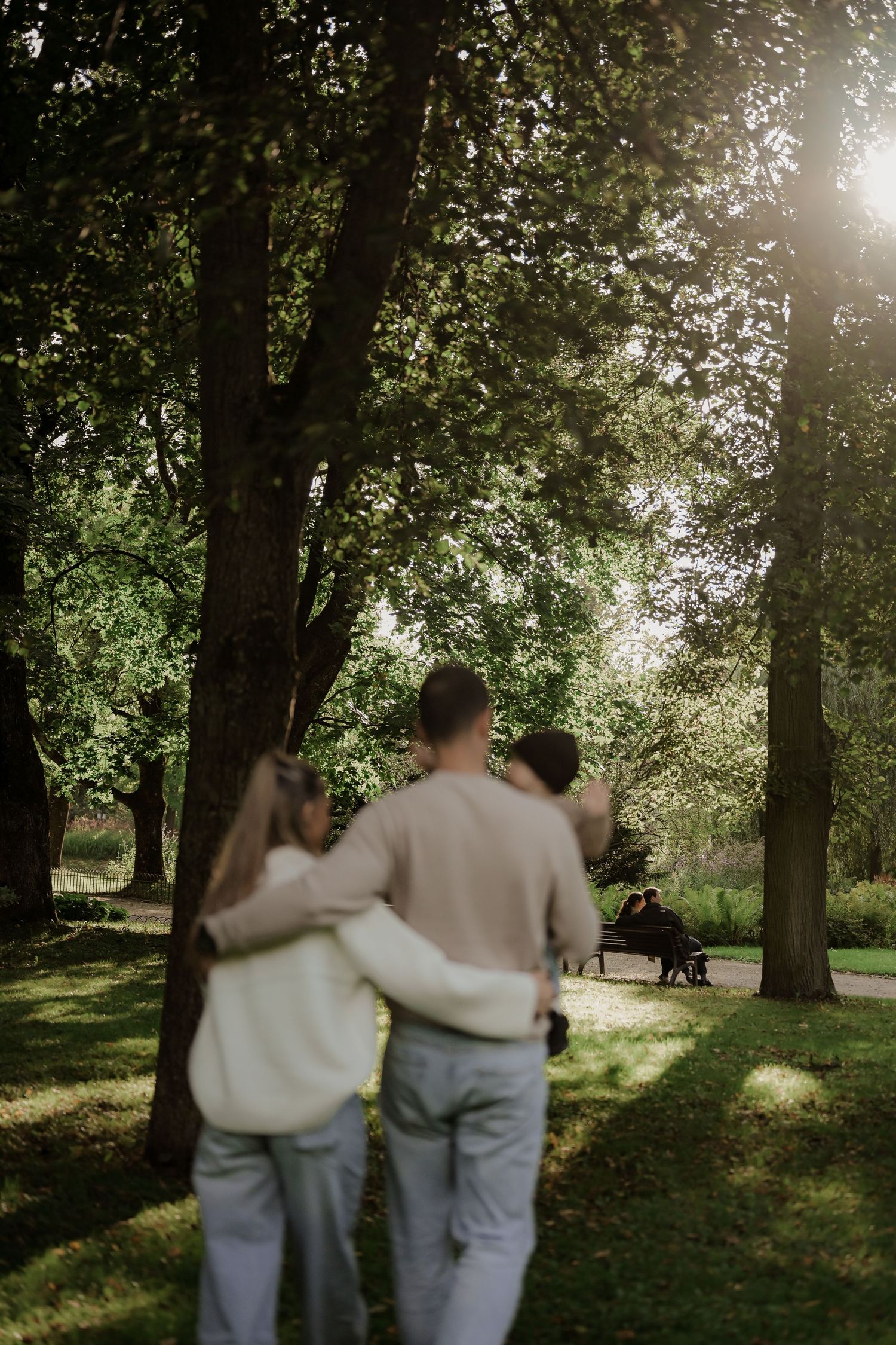 A couple walking arm in arm through a sunlit park with tall trees and green grass.
