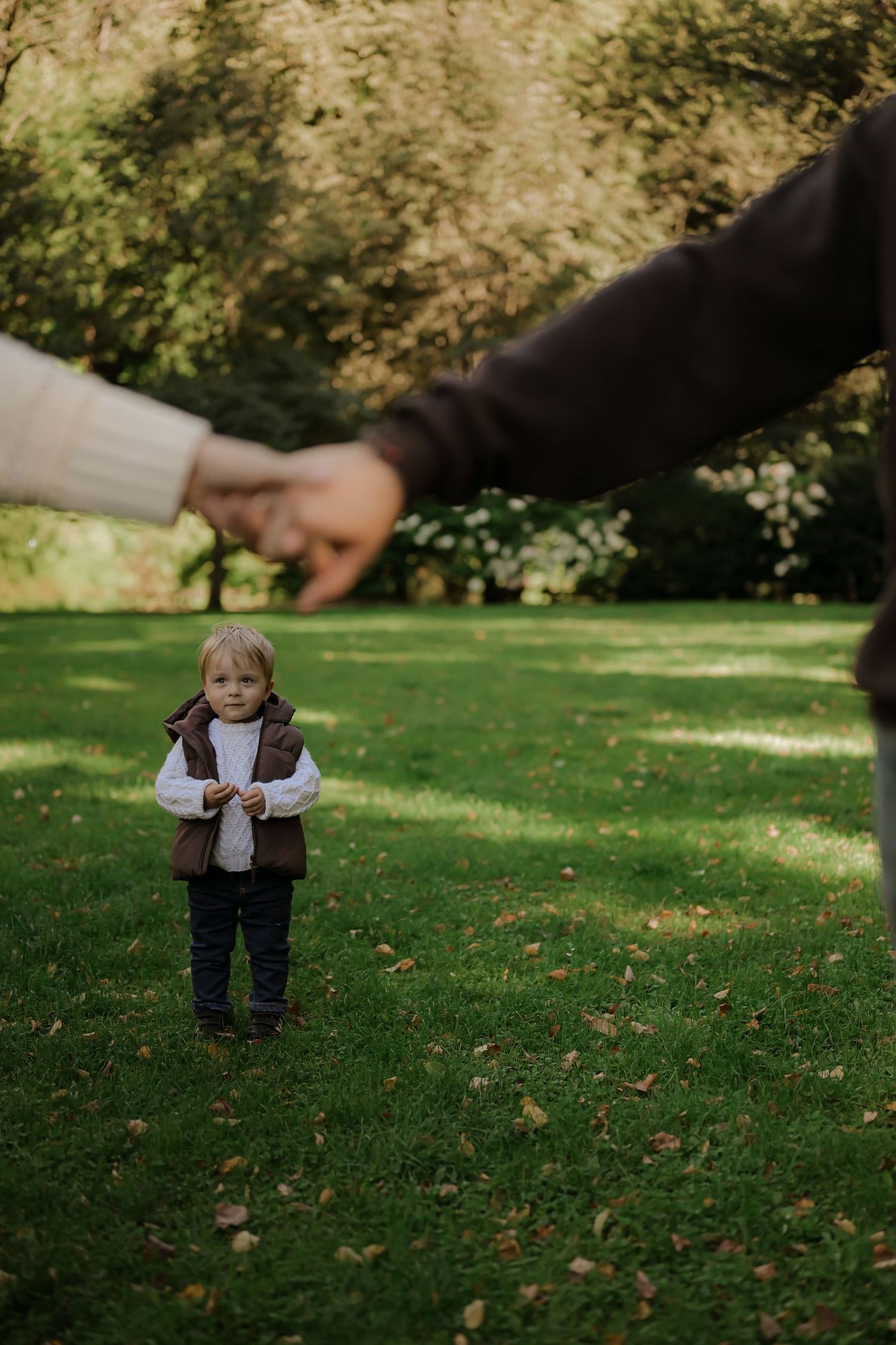Toddler in brown vest stands on green grass while parents hold hands in the foreground of a lush park.