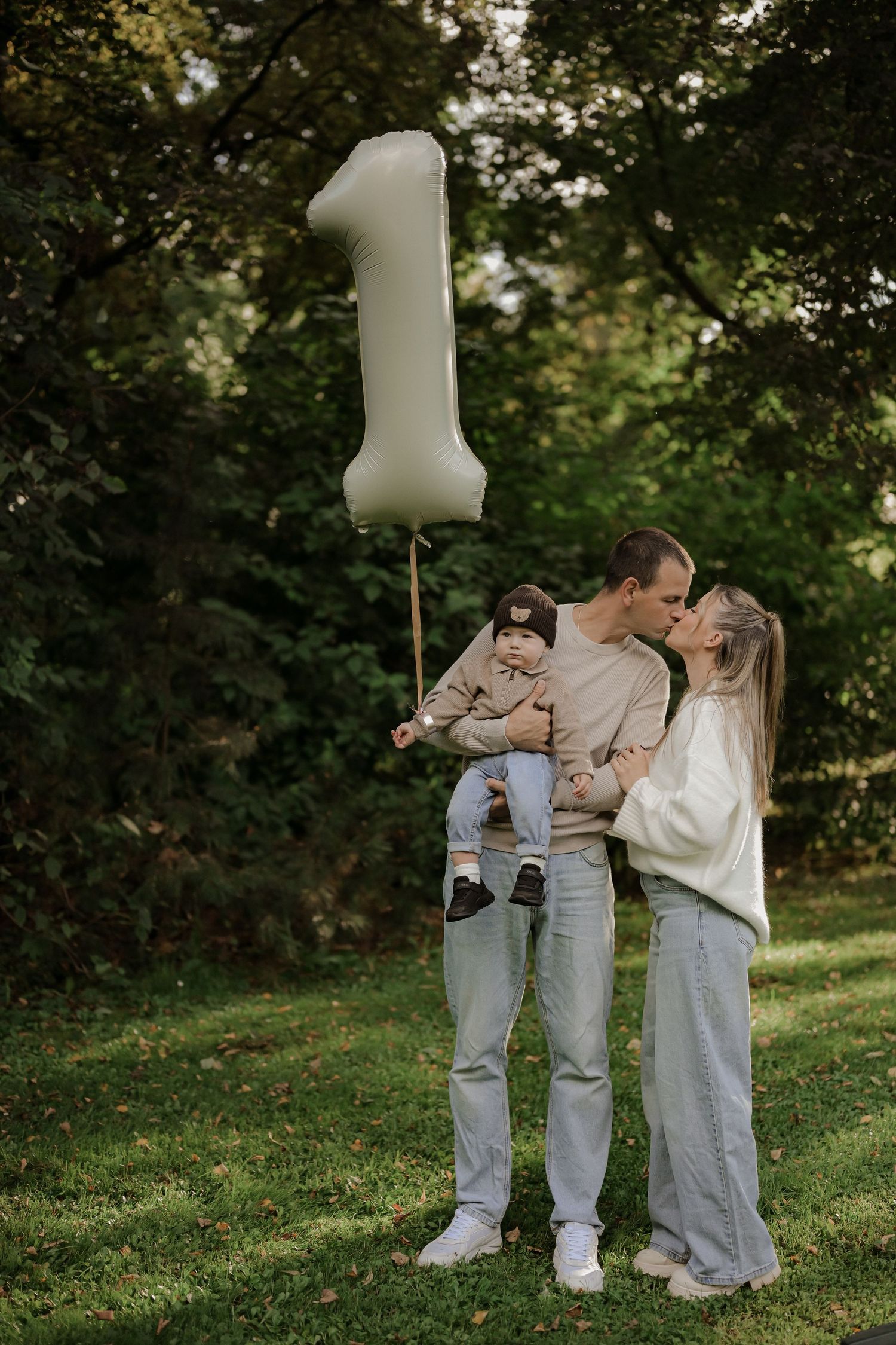 A couple celebrates their baby's first birthday in a park, holding a large number 1 balloon while kissing.