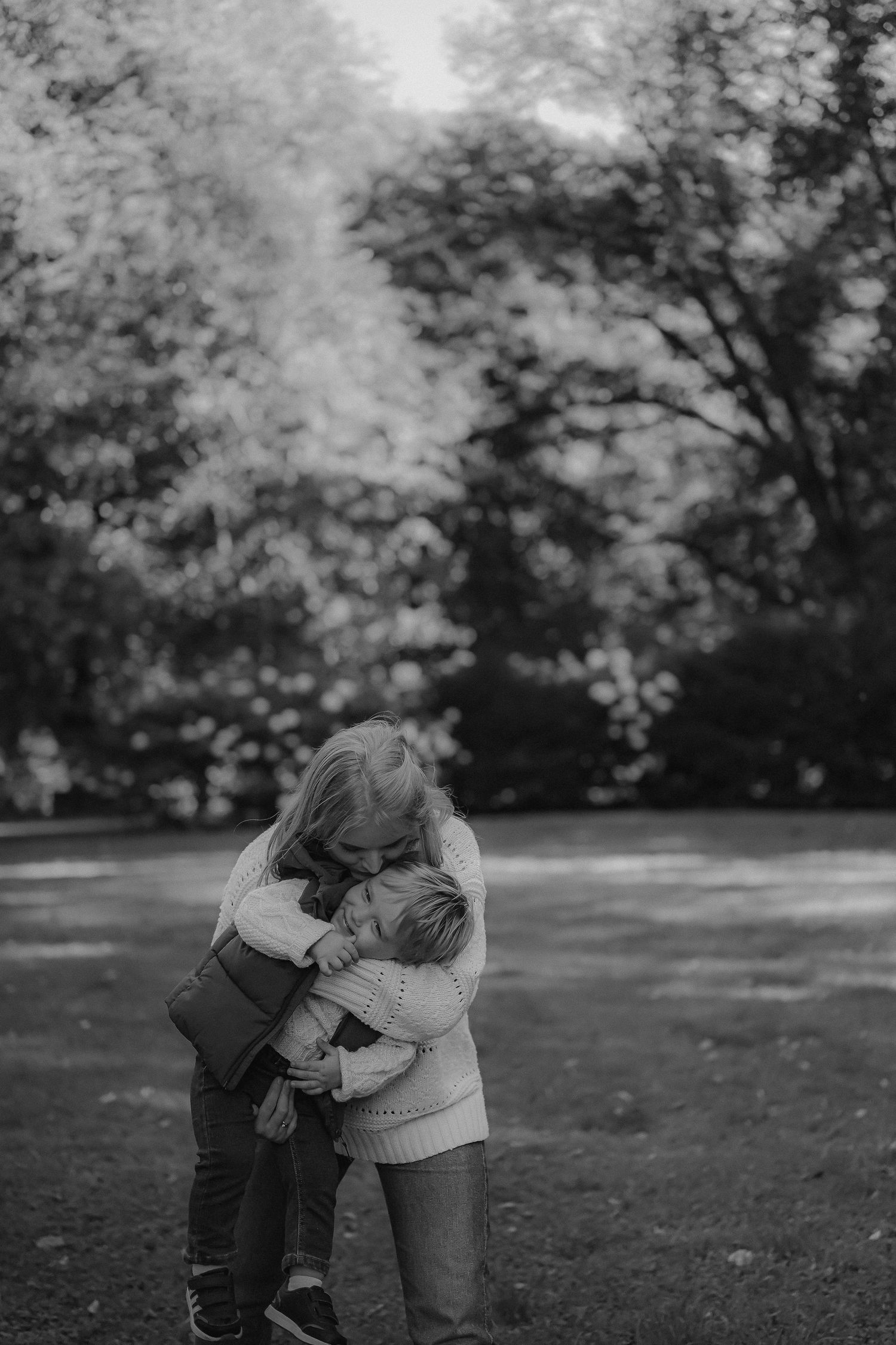 A mother and child share a tender embrace in a black and white outdoor park photo.