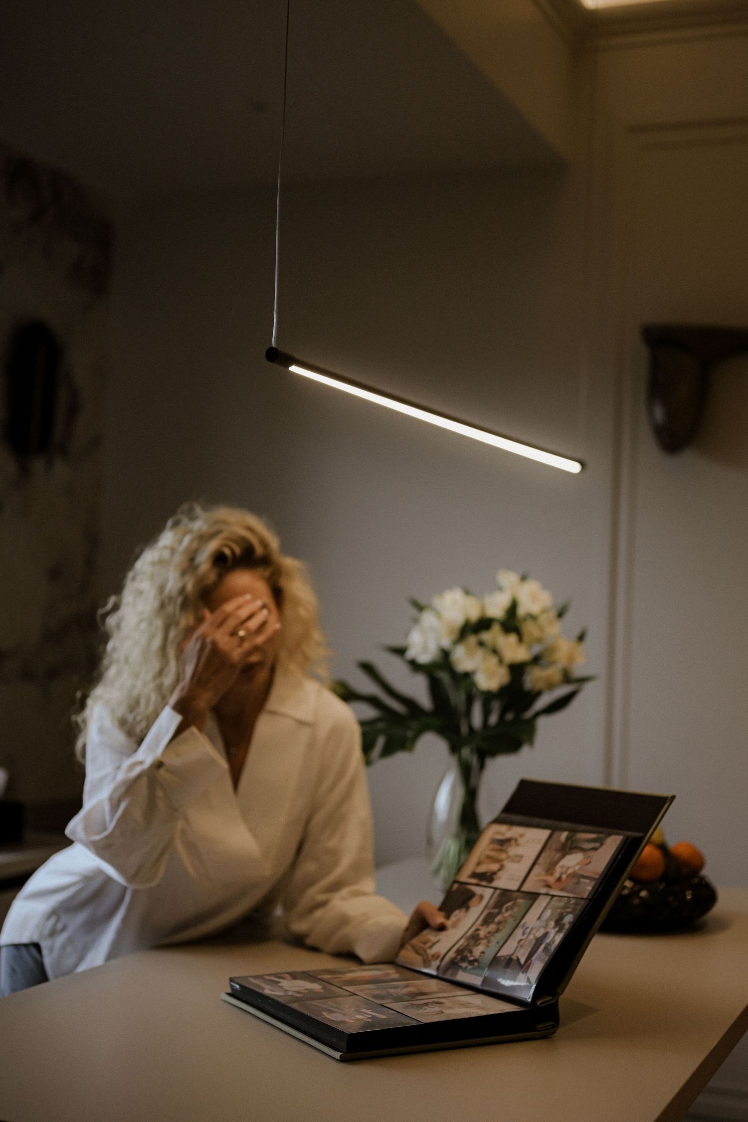 Woman with curly blonde hair sitting at a table looking at a photo album under a modern pendant light.