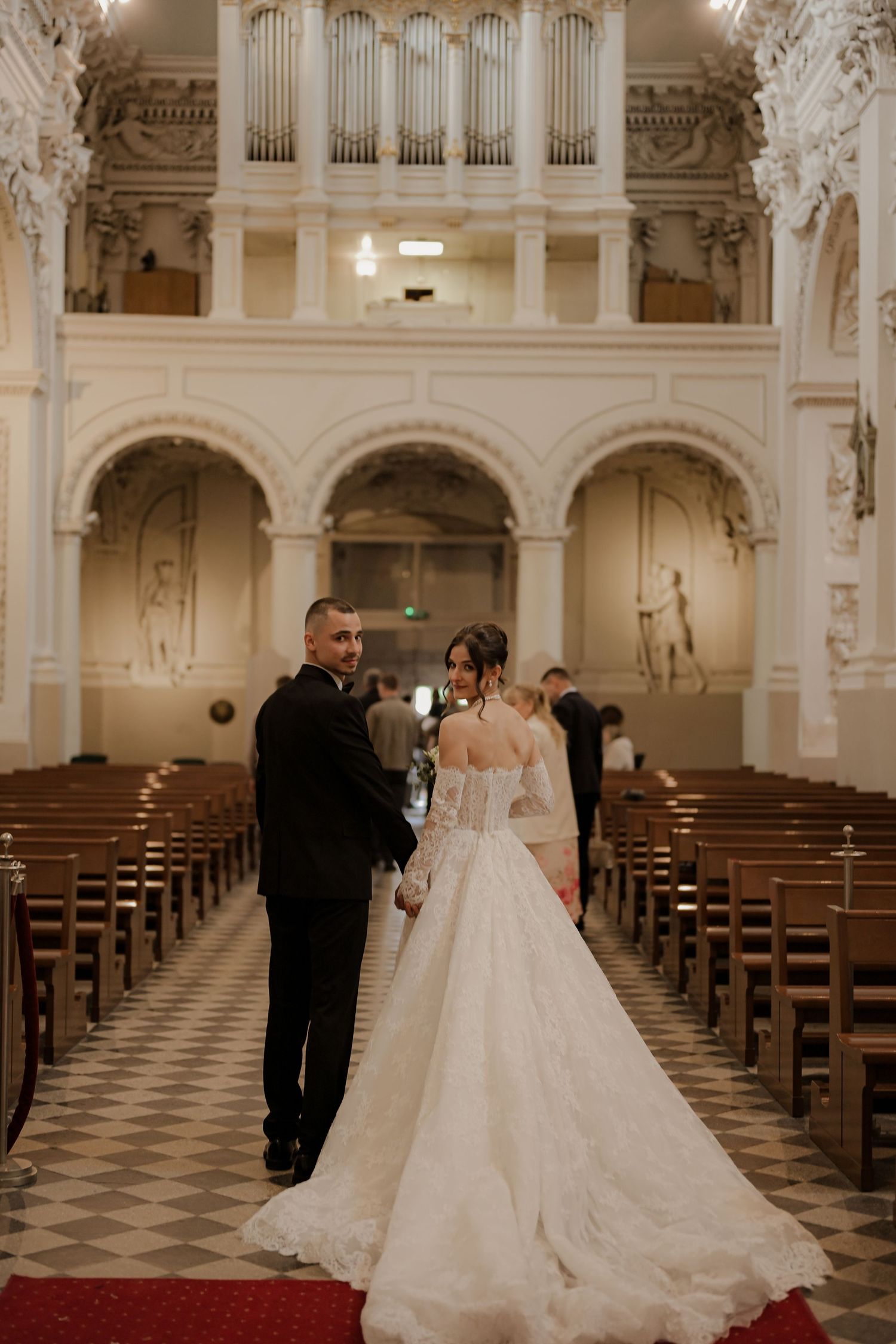 Bride and groom walk down the aisle of an ornate white church with pipe organ and wooden pews.