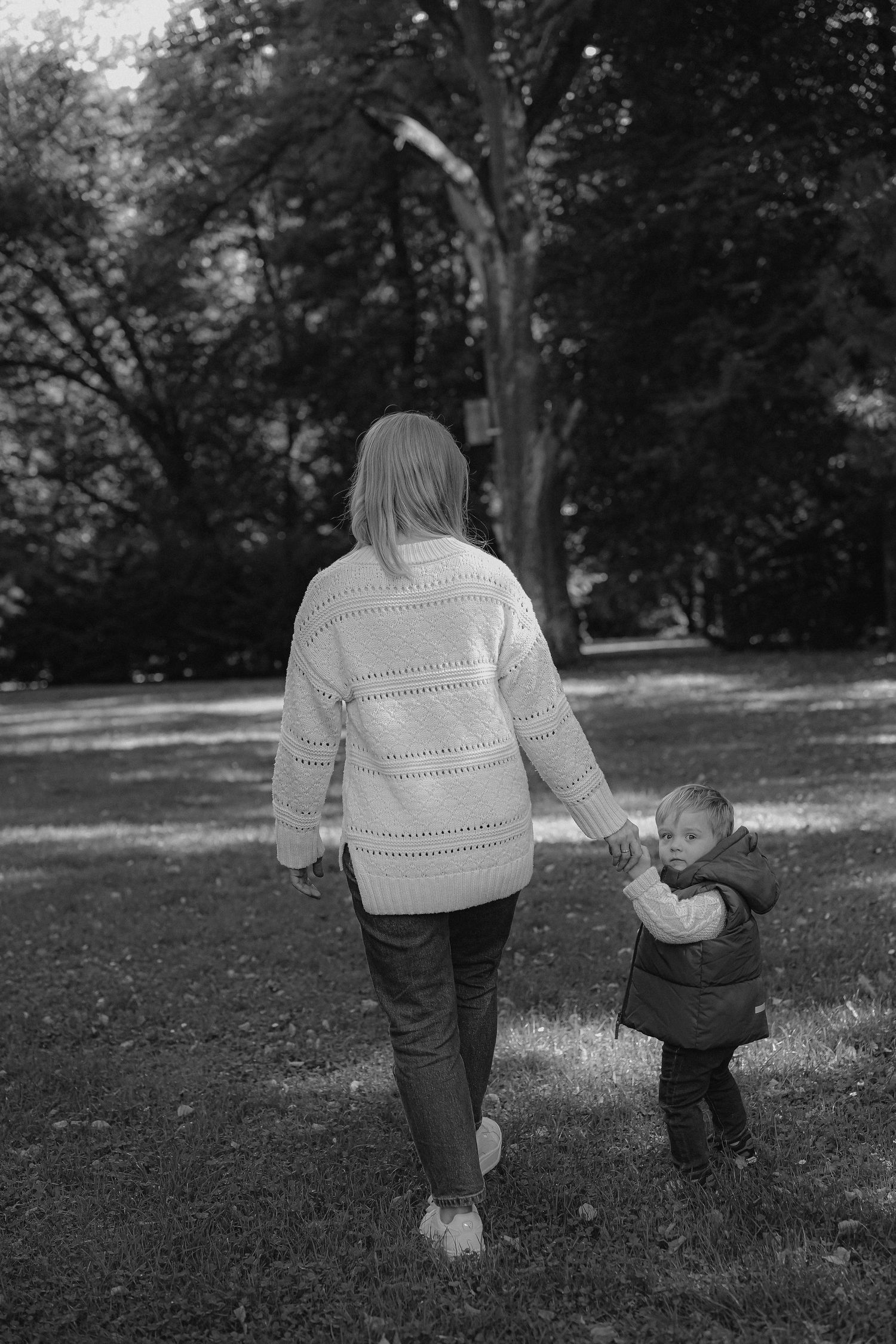 Black and white photo of a mother and toddler walking hand in hand through a lush green park.