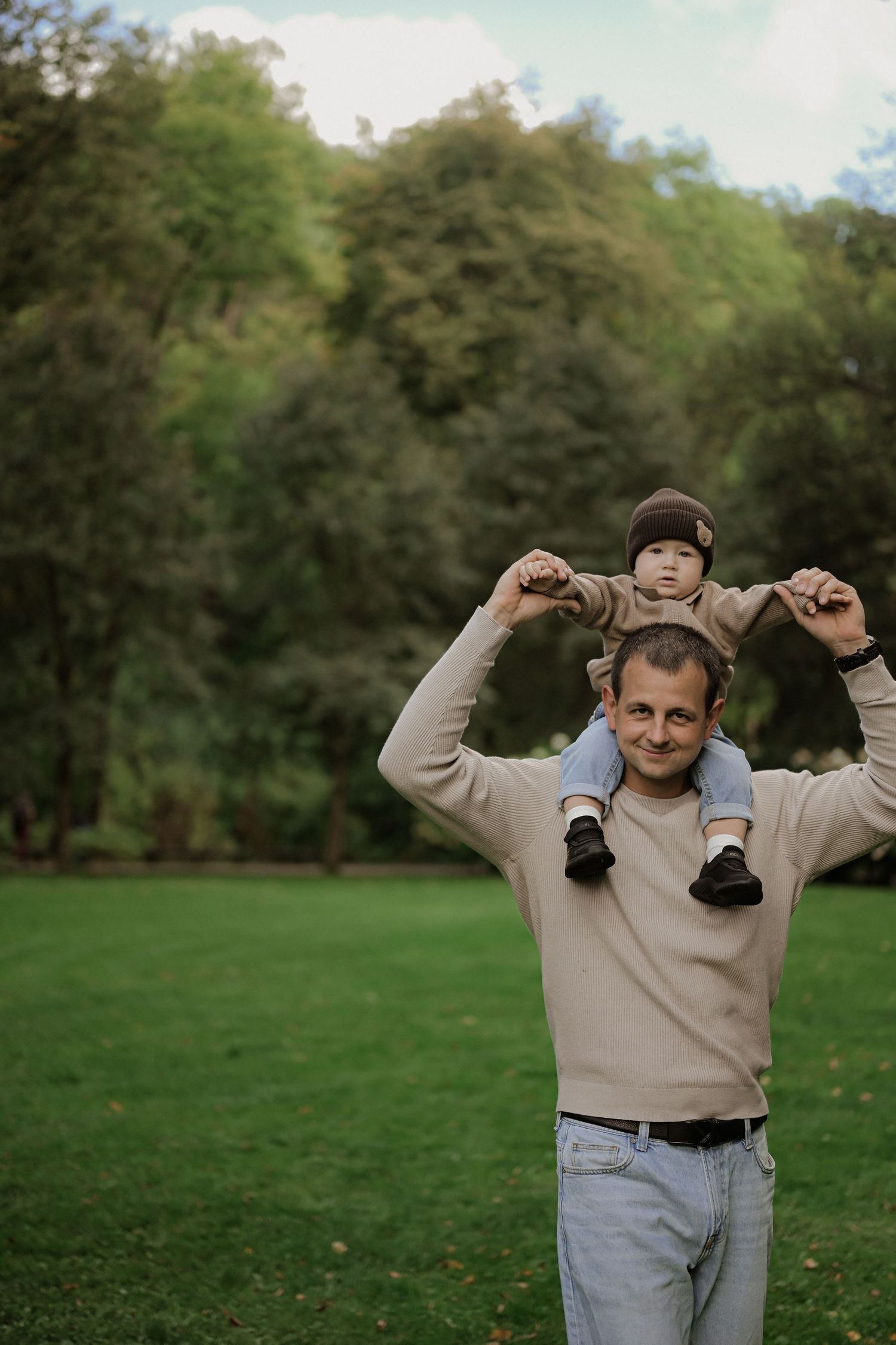 A smiling father carrying his toddler on his shoulders in a lush green park setting.
