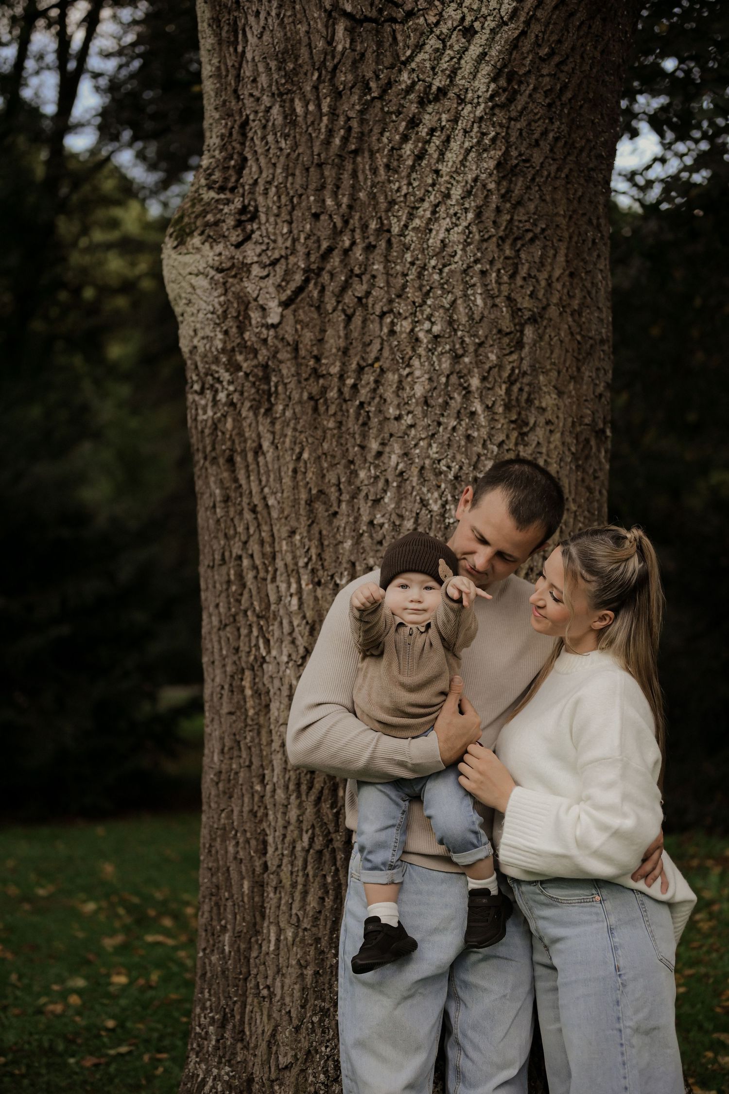 Young family with baby poses by large tree trunk in park, parents smiling at child in autumn setting.