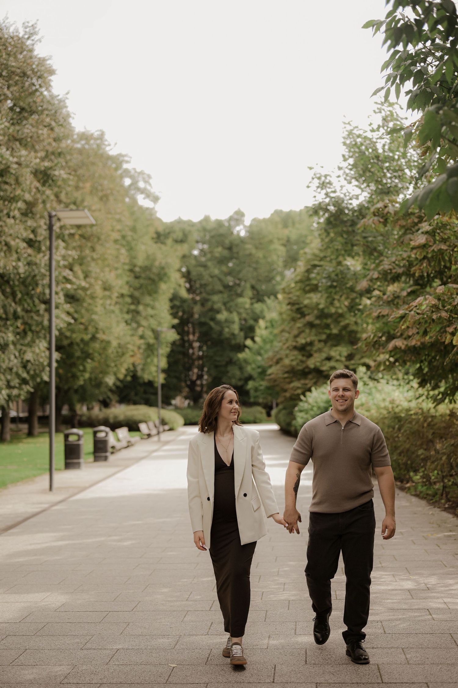 Couple holding hands and walking together on a tree-lined park path, woman in white blazer, man in brown polo shirt.