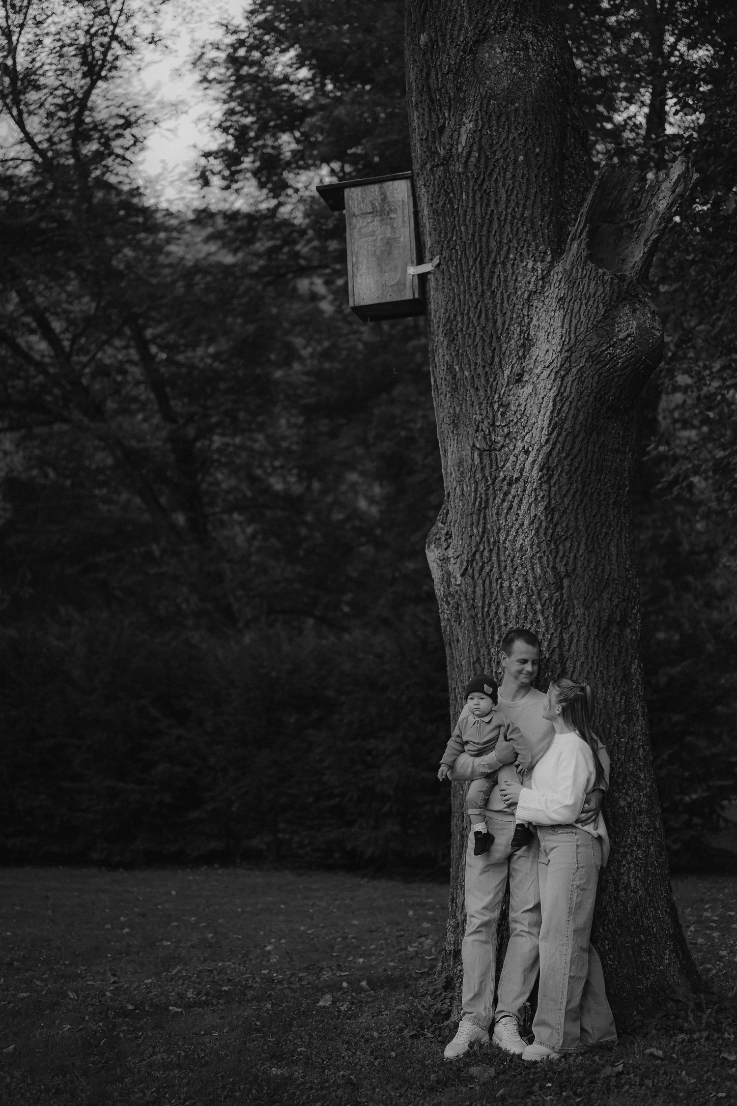 Black and white photo of a family with a baby leaning against a large tree in a lush park setting.