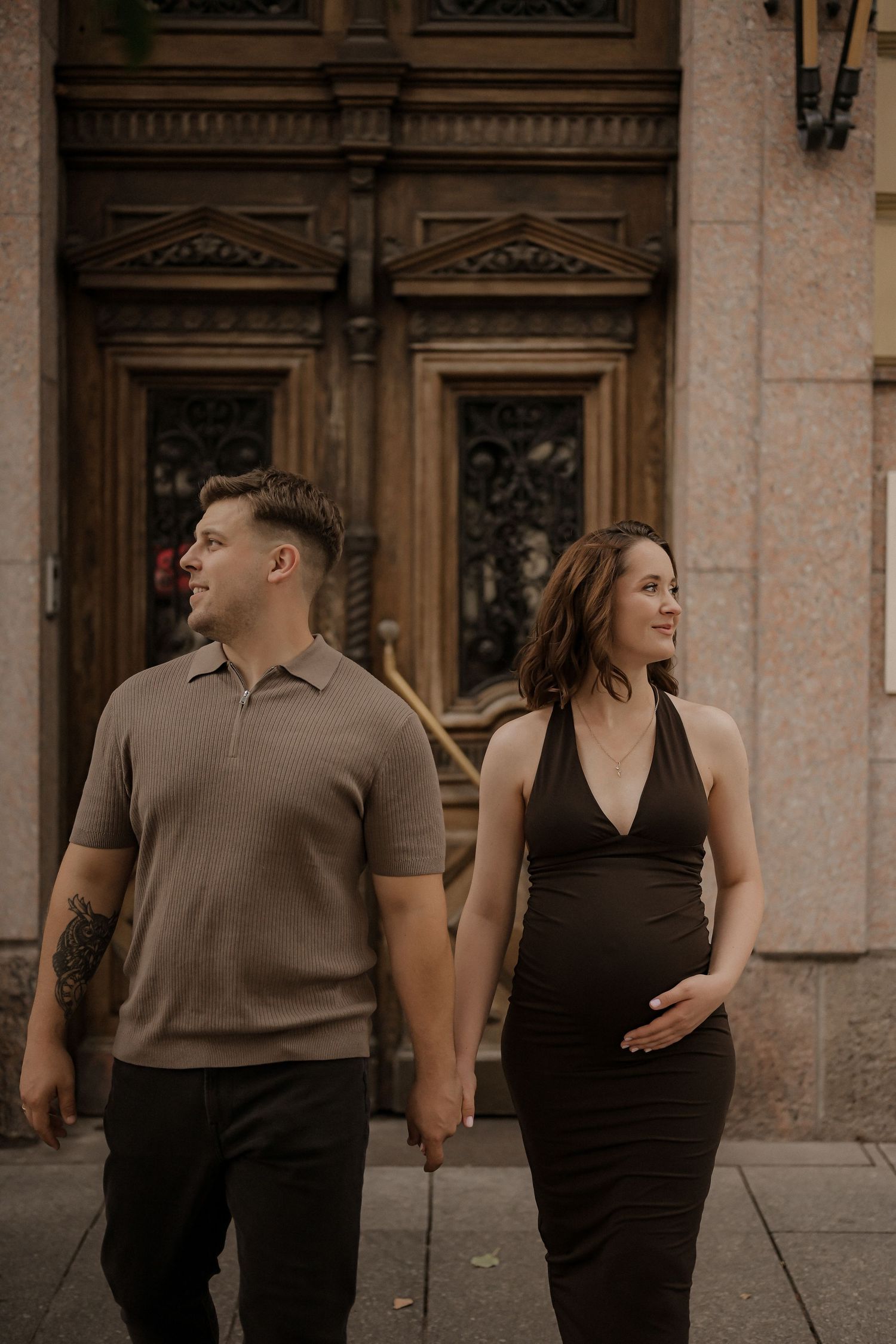 Couple walking hand-in-hand in front of ornate wooden doors, woman in black halter maternity dress, man in tan polo shirt.
