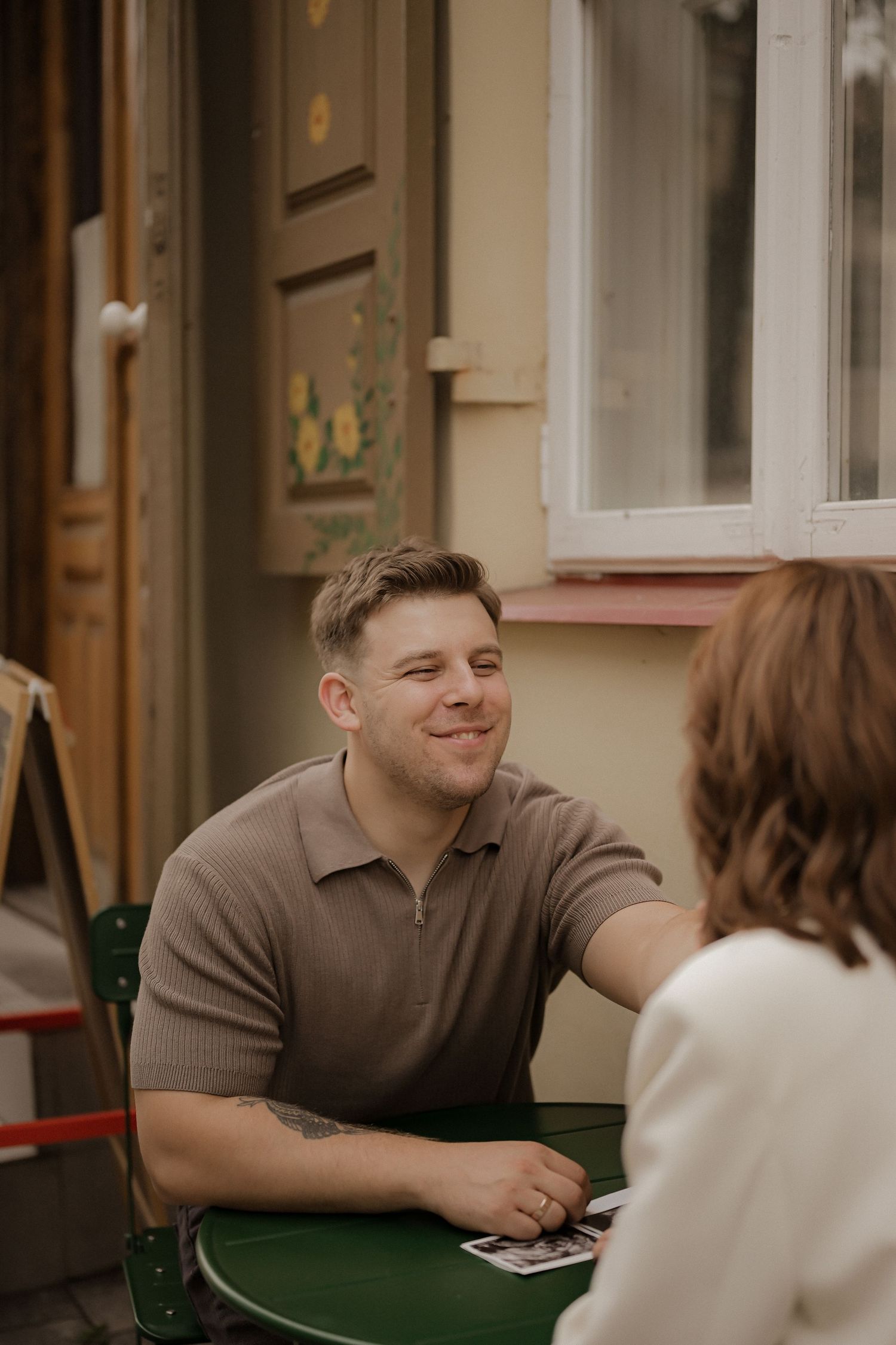 A smiling man in a brown polo shirt sits at a green outdoor cafe table, engaged in conversation with a woman.