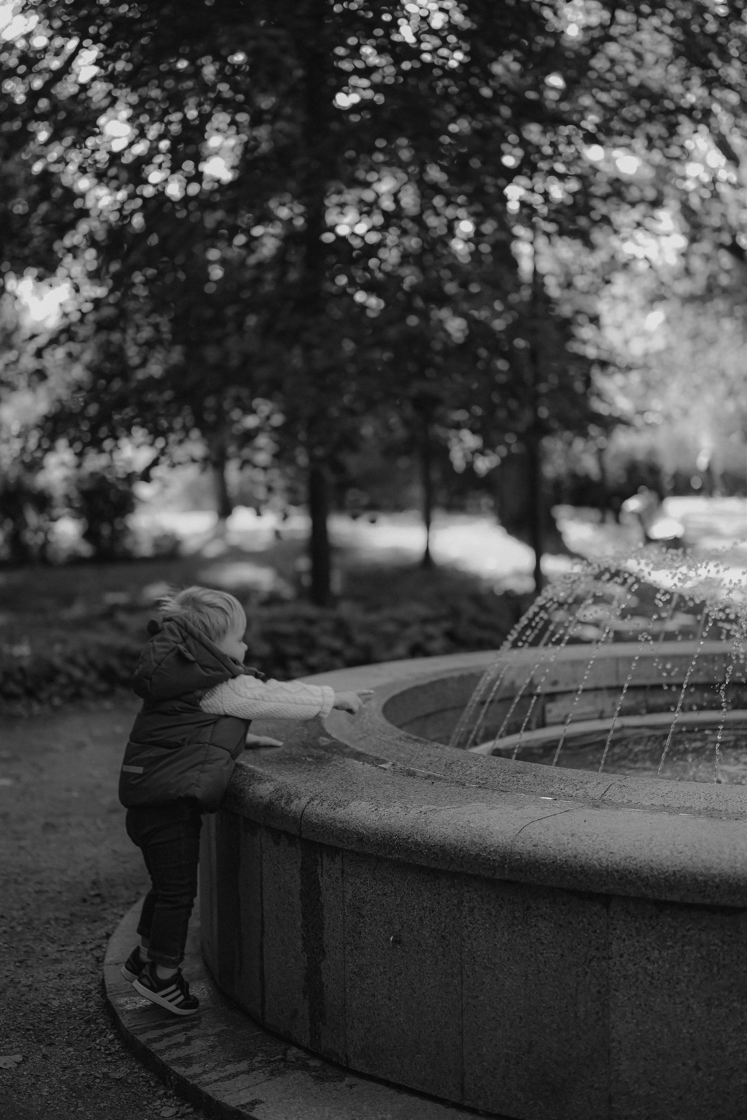 Black and white photo of a young child reaching into a park fountain surrounded by trees.