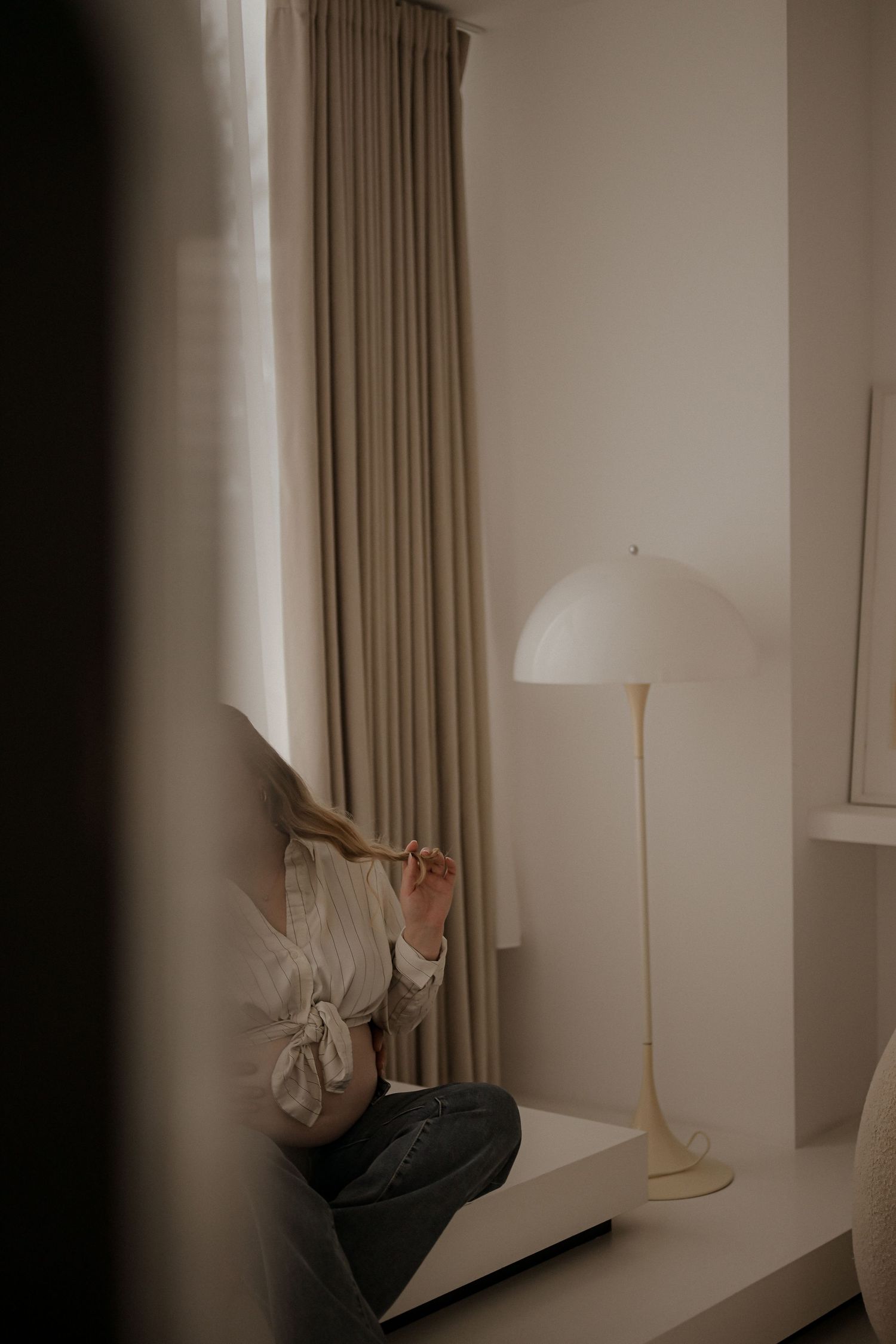 Person sitting by a window with beige curtains next to a white floor lamp in a minimalist room.