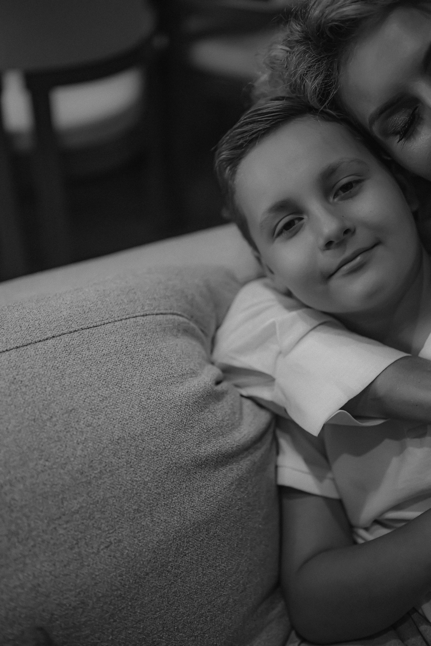 Black and white photo of a young boy resting his chin on a sofa arm, being embraced by a woman behind him.