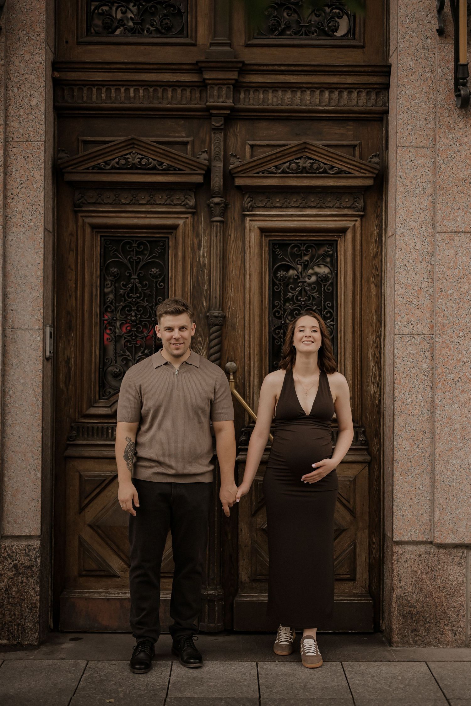 A couple holds hands in front of an ornate wooden door, the woman visibly pregnant.