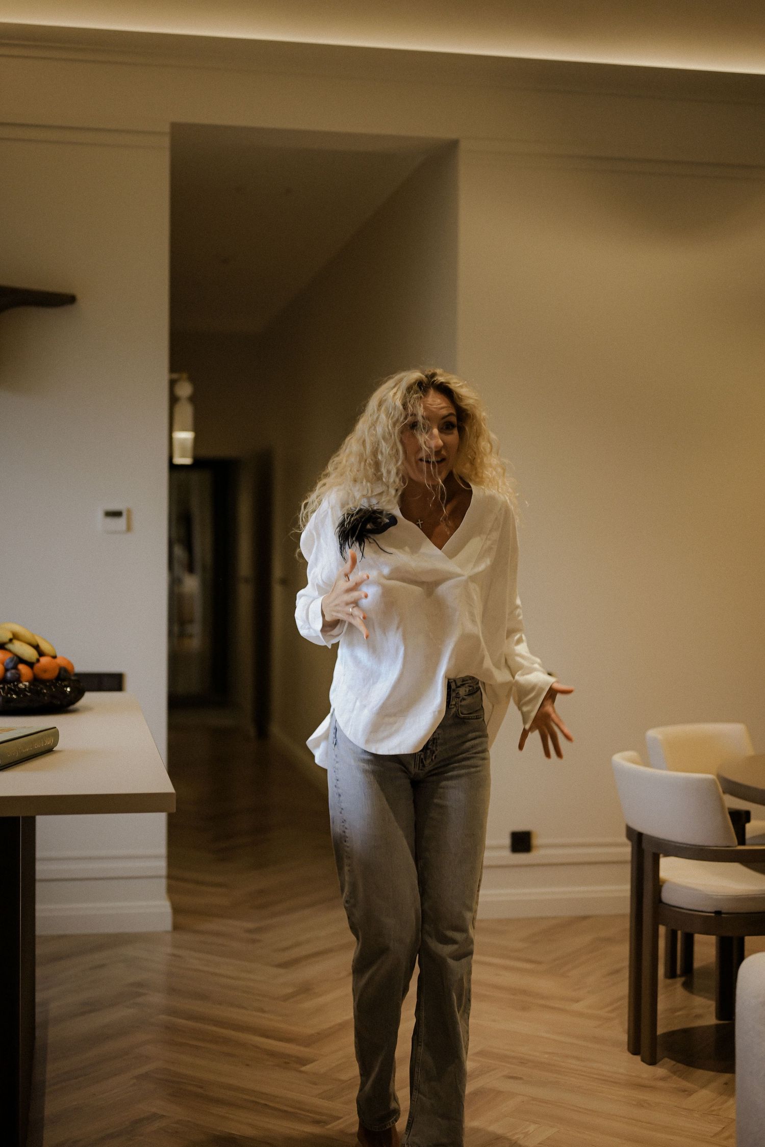 Woman in white blouse and gray jeans walking through modern apartment interior with herringbone wood floors.