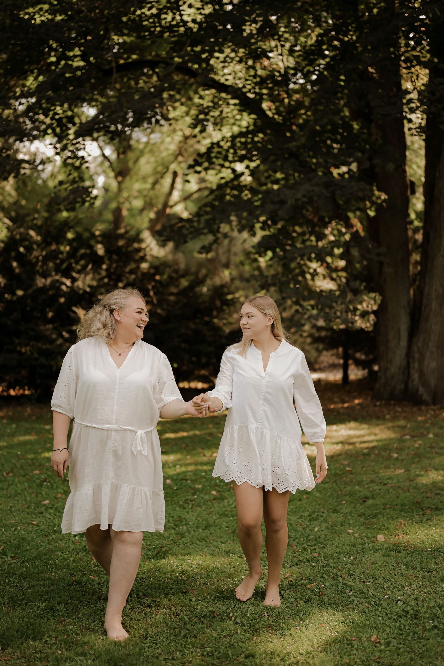 Two women in white dresses hold hands and smile while walking barefoot on green grass in a sunlit park.
