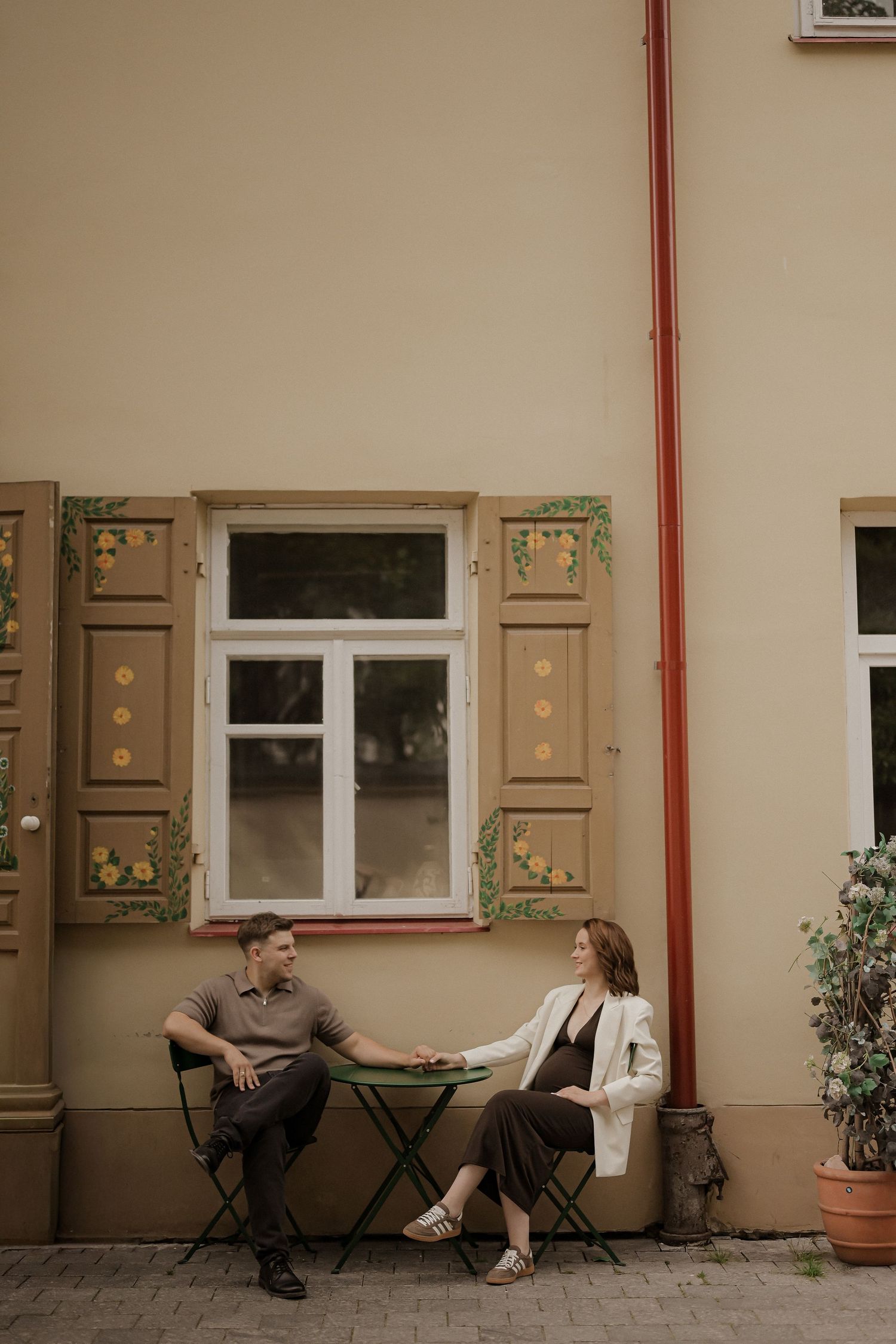 Expecting couple sitting at a small table outside a charming building with decorative painted shutters.
