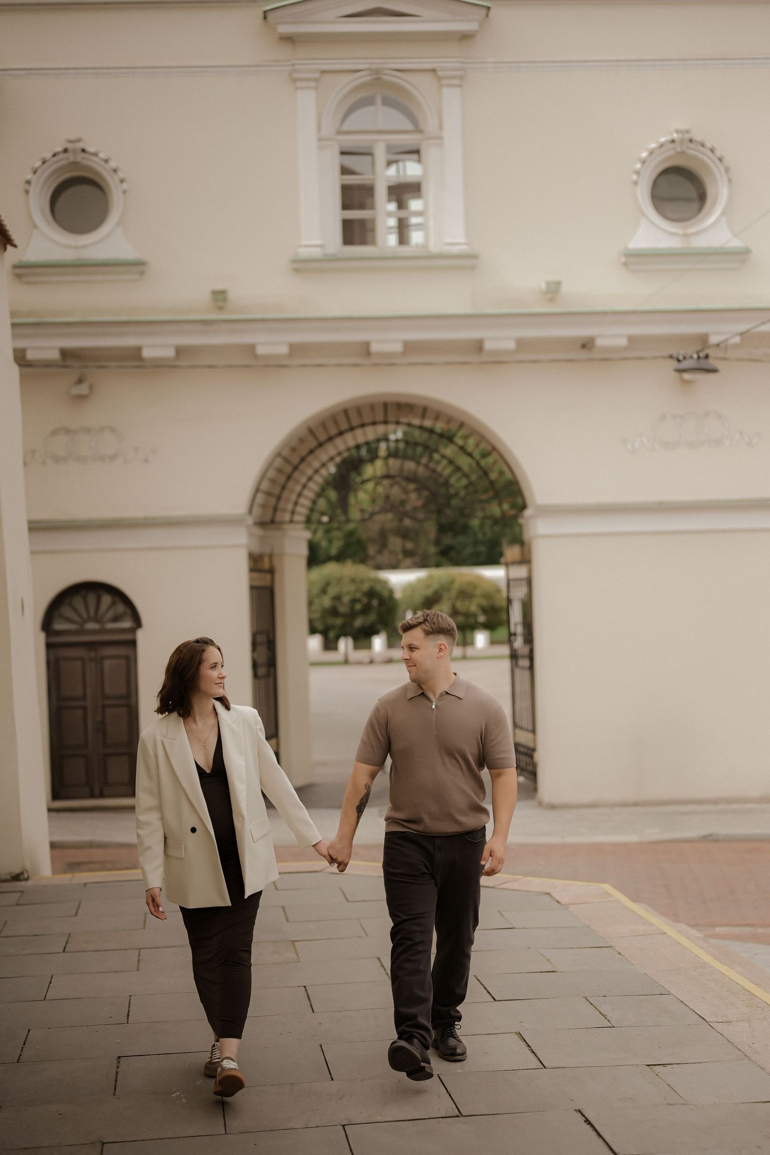 A couple walks hand in hand past a classical white building with an arched gateway.