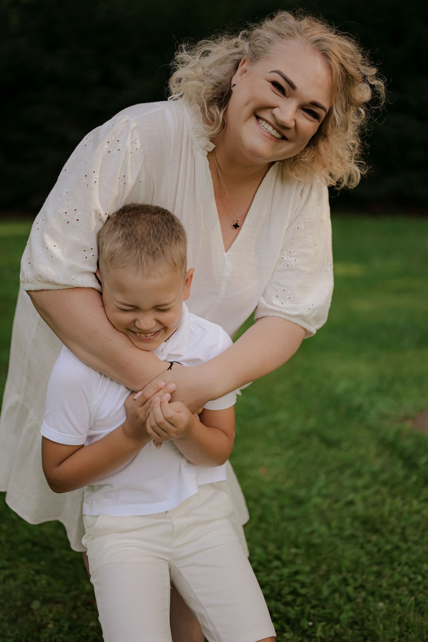 A smiling woman in white hugs a laughing boy outdoors on green grass, both wearing white clothing.