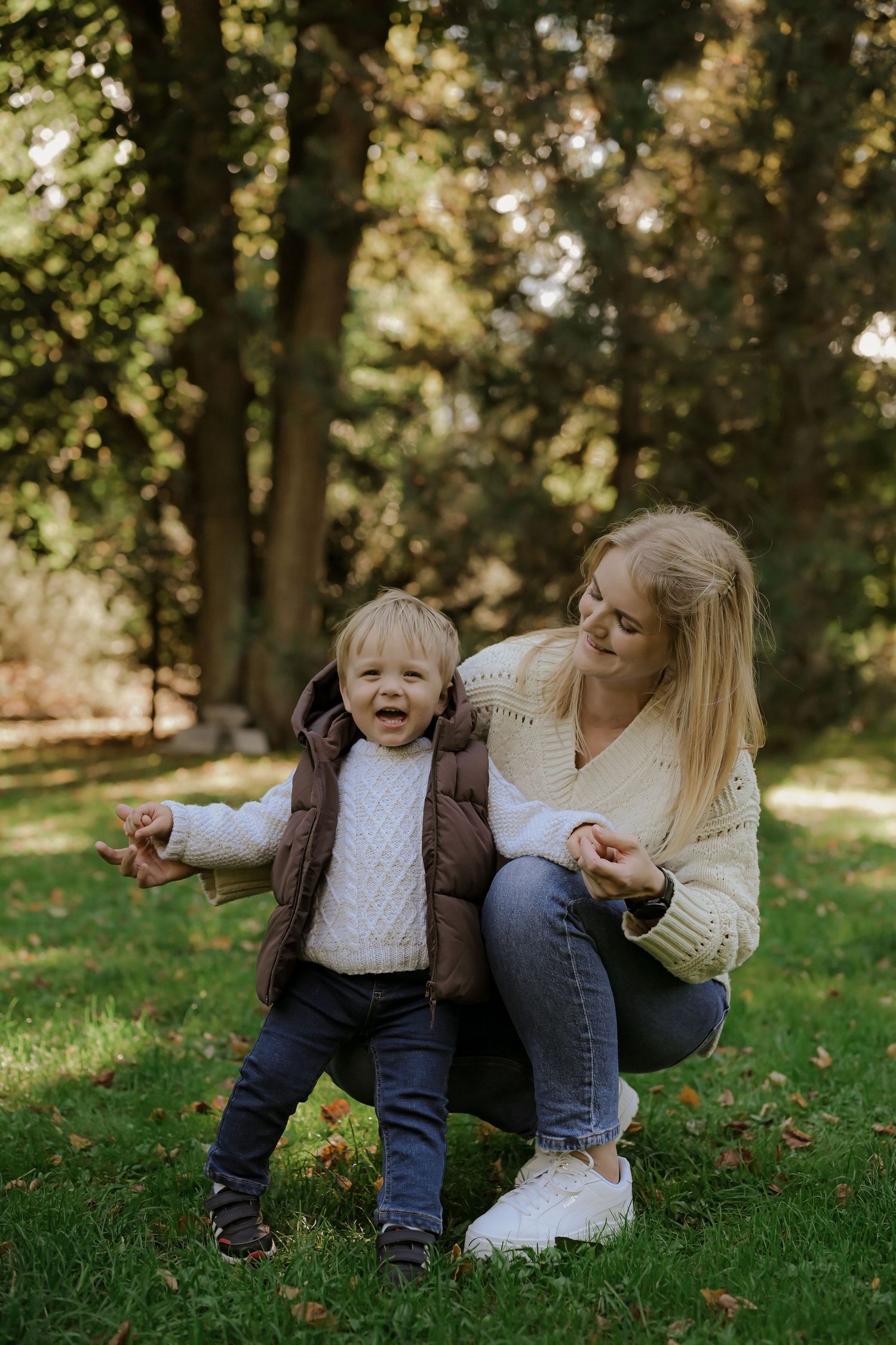 A smiling blonde mother crouches beside her laughing toddler in a brown vest on green grass in a park.