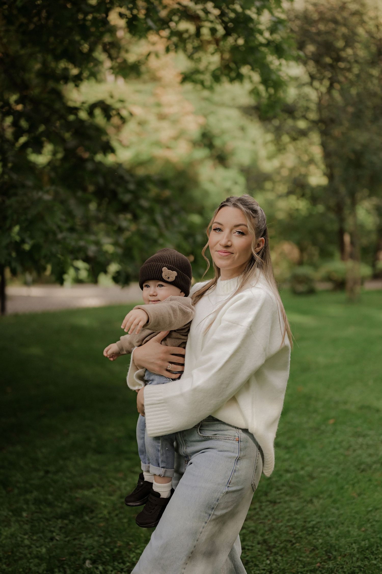 Young woman in cream sweater and jeans holds a baby in a brown bear hat in a lush green park.