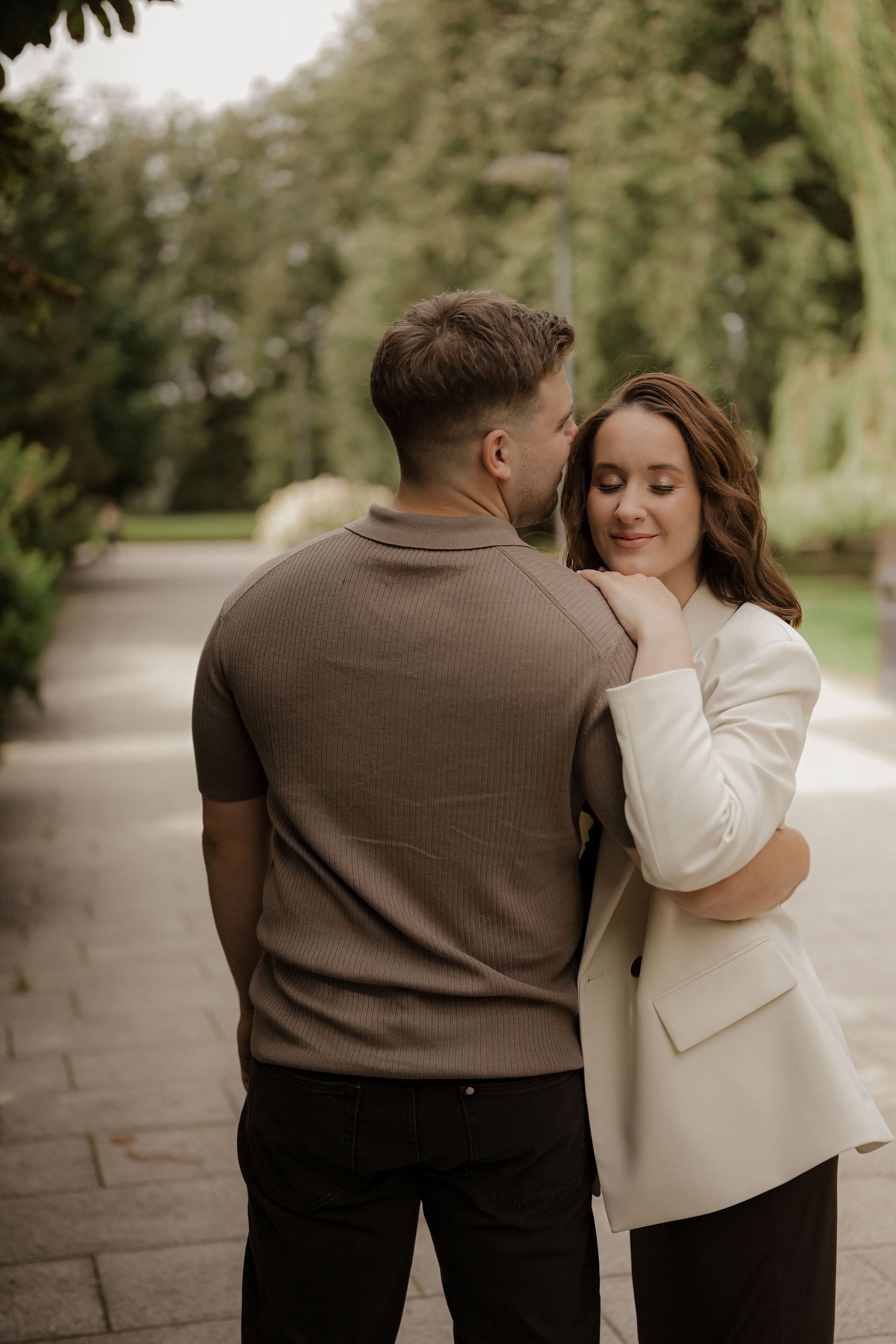 Romantic couple embracing on a park pathway, woman in white blazer resting against man in brown polo shirt.