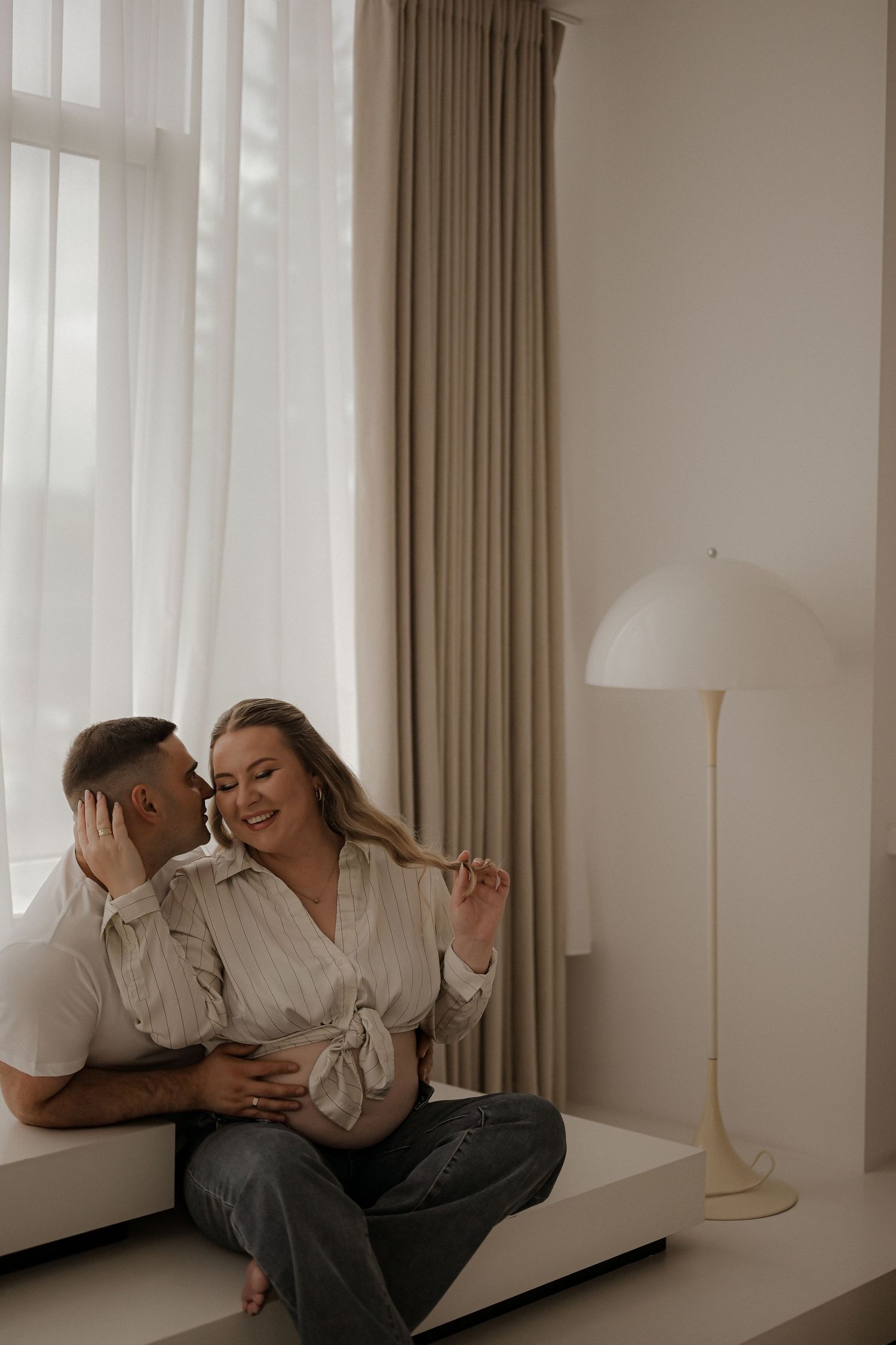 Pregnant couple sitting together on a white bench by curtained window, sharing a tender moment in a bright minimalist room.