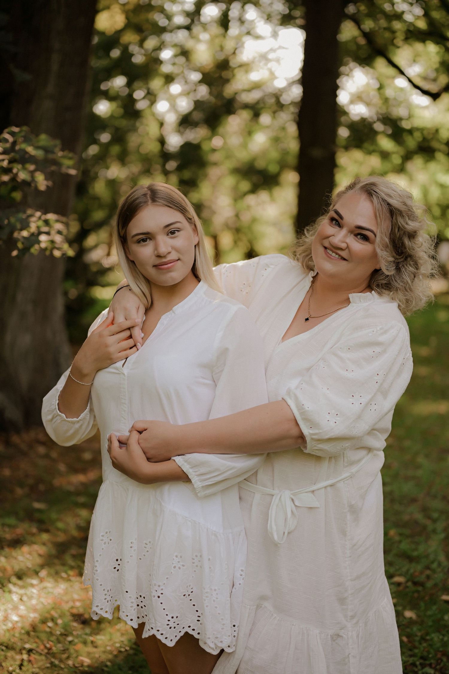 Two women in white dresses embrace warmly outdoors in a lush green park setting.
