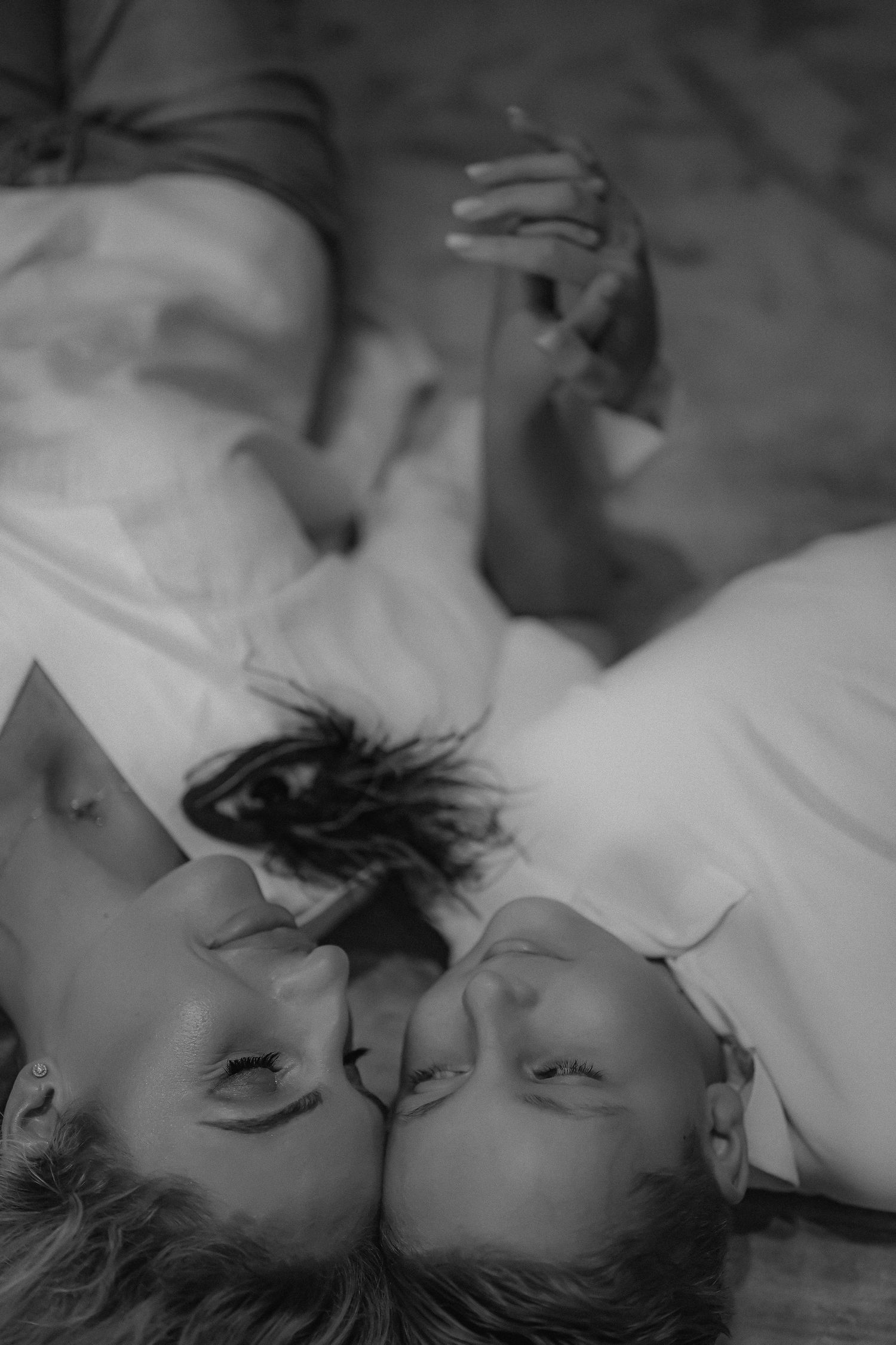 Black and white photo of two people lying down face-to-face, smiling and holding hands in a romantic moment.