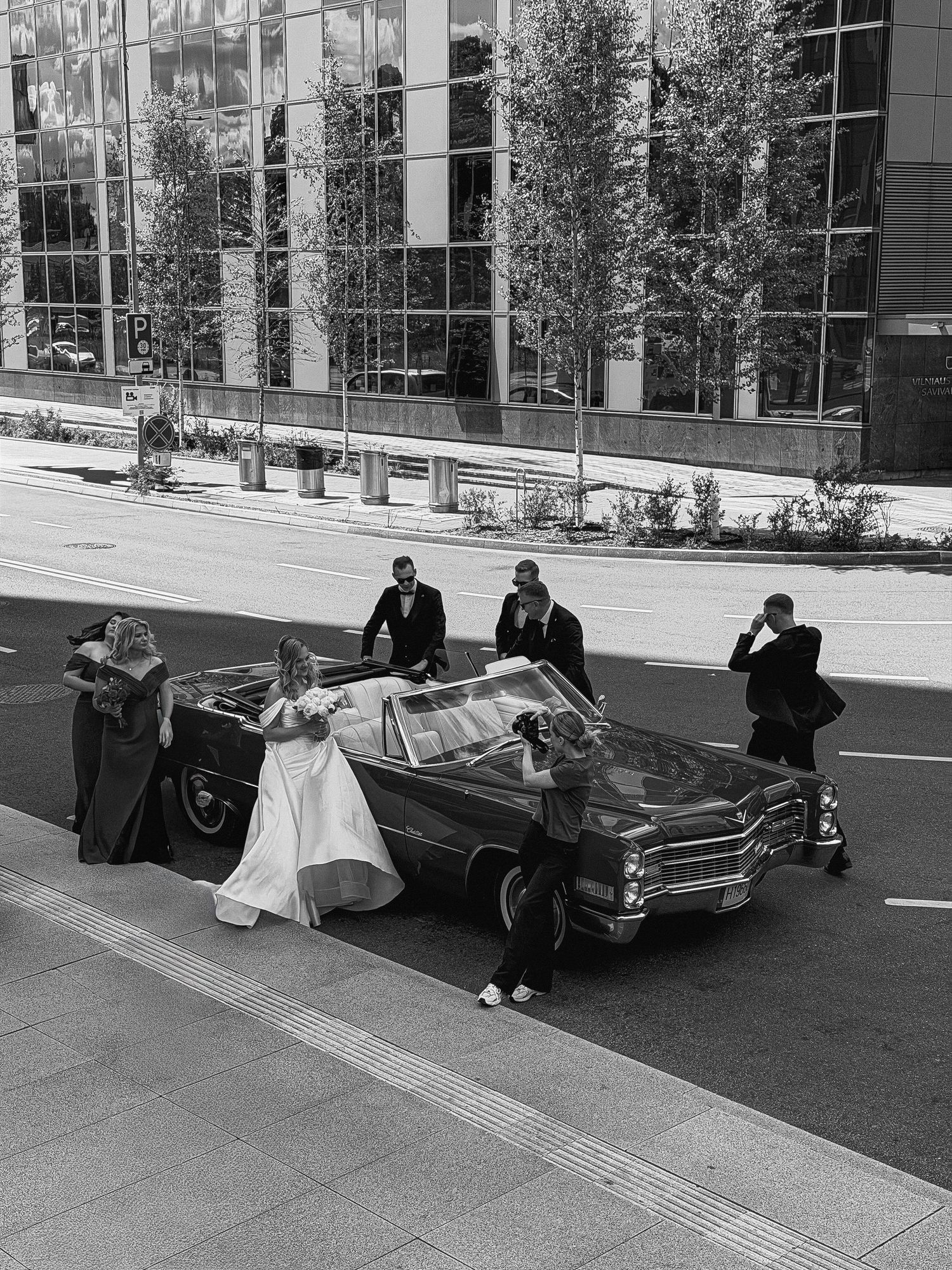 Black and white photo of a wedding party posing around a vintage convertible car on a city street.