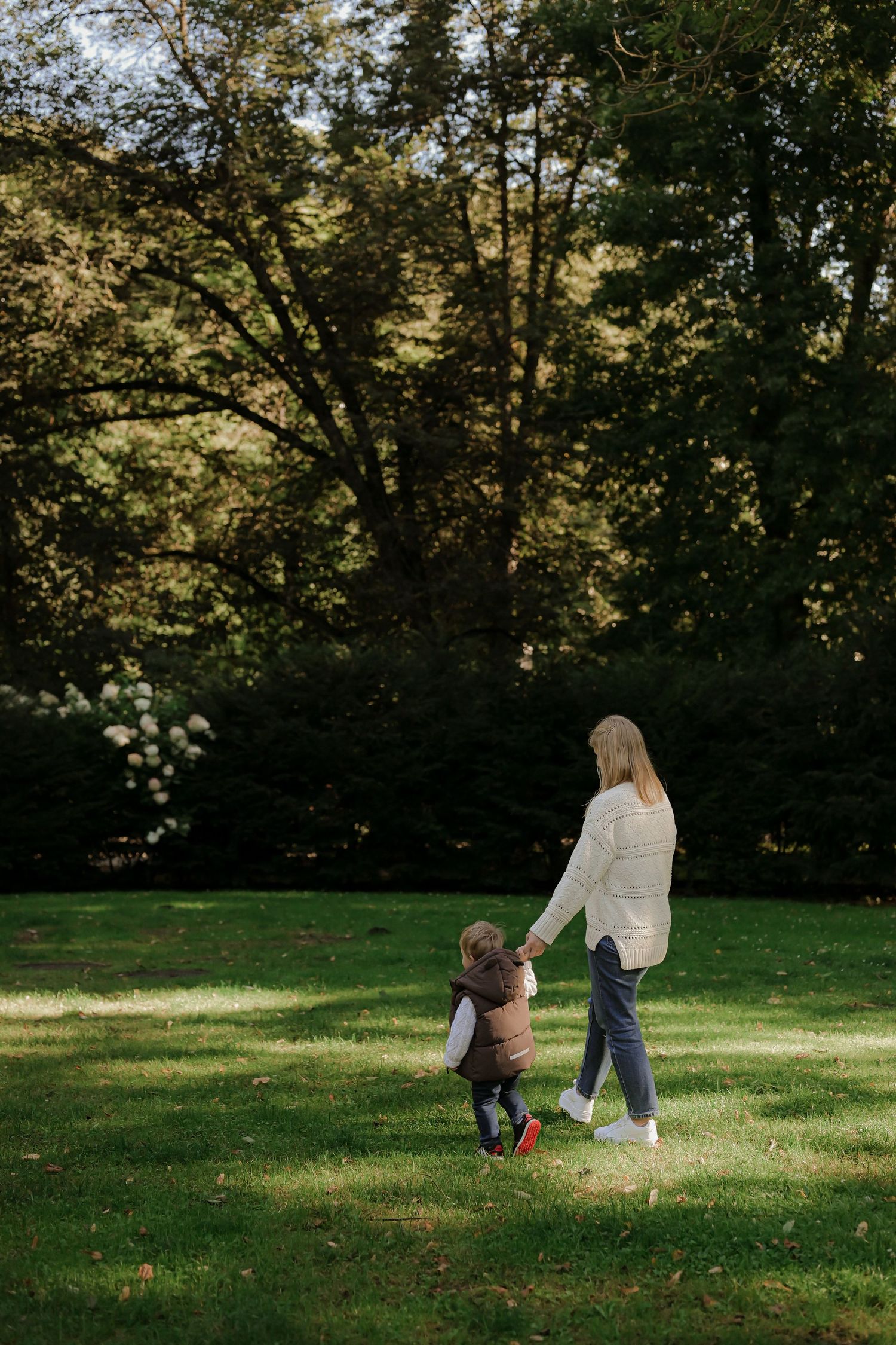 A blonde mother holds her toddlers hand while walking across a sunny green park lawn surrounded by trees.