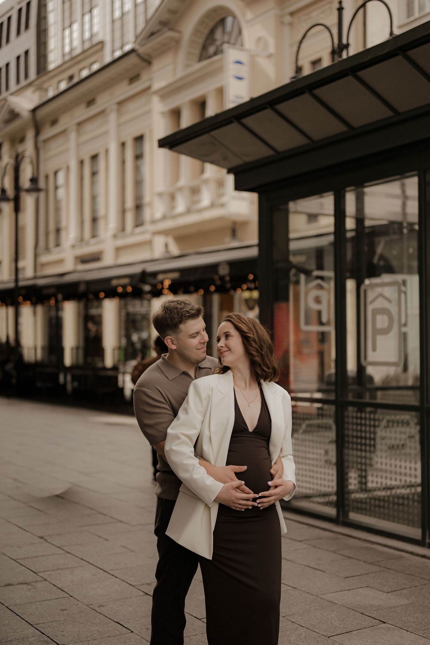 Pregnant woman in white blazer embraced by partner on a city street with elegant architecture.