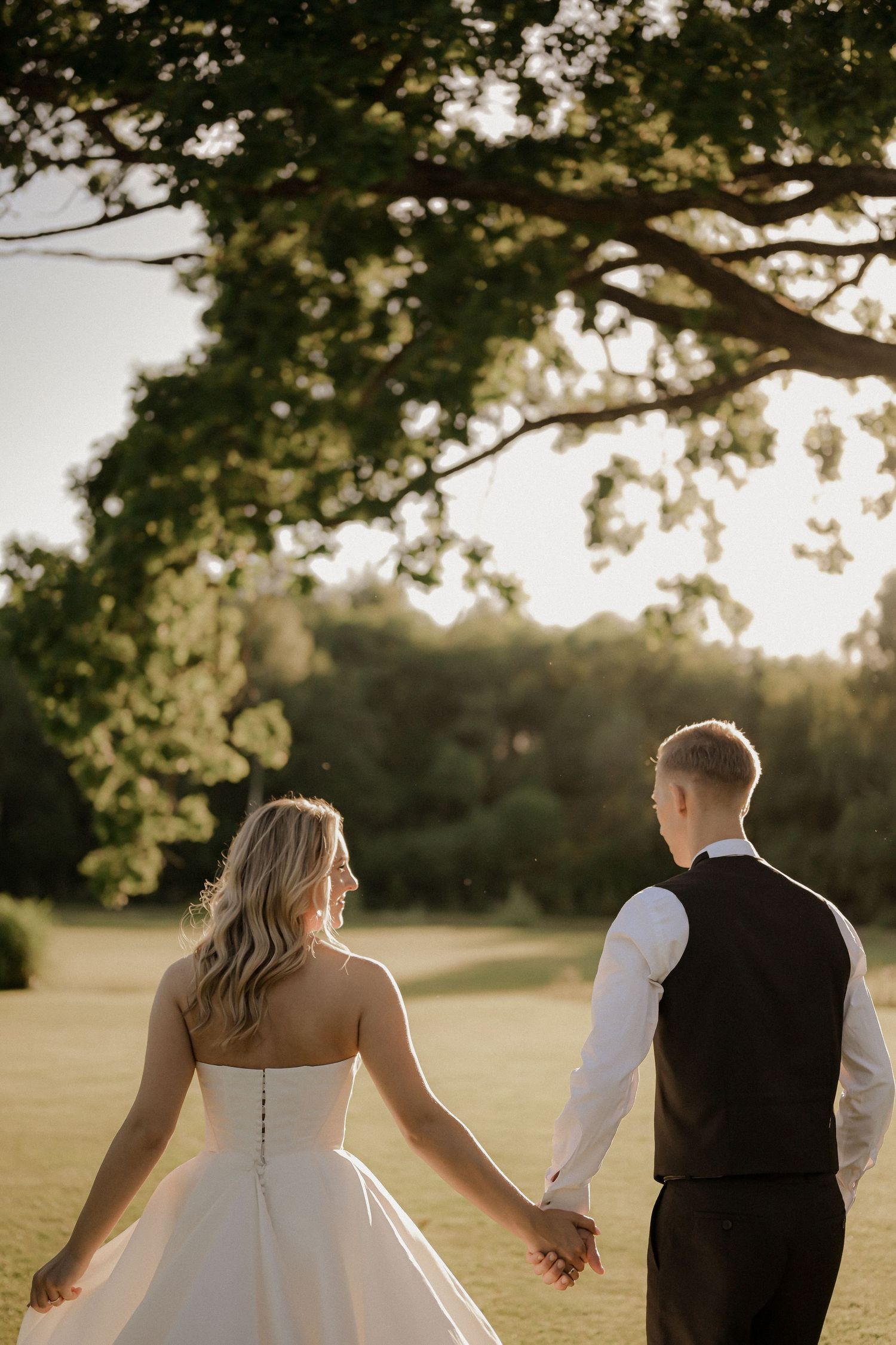 Bride and groom holding hands walking through lush green garden at golden hour, bride in strapless gown.