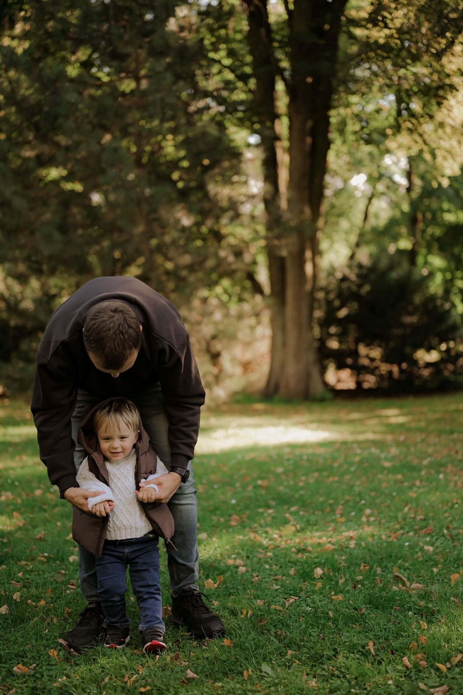 Father bending down to help his toddler walk on green grass in a sunny autumn park.