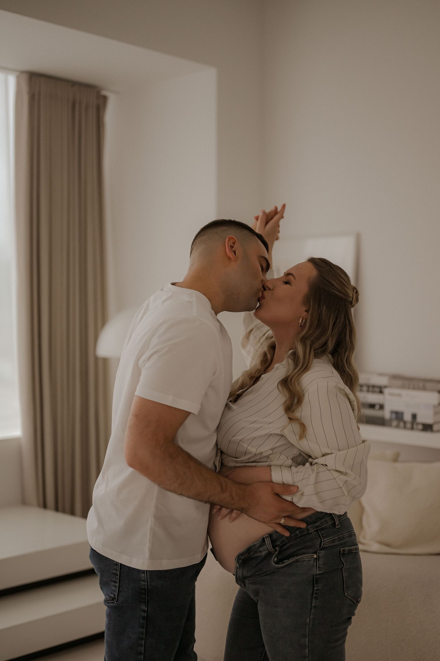 Couple sharing a tender kiss while cradling a pregnant belly in a bright, minimalist living room.