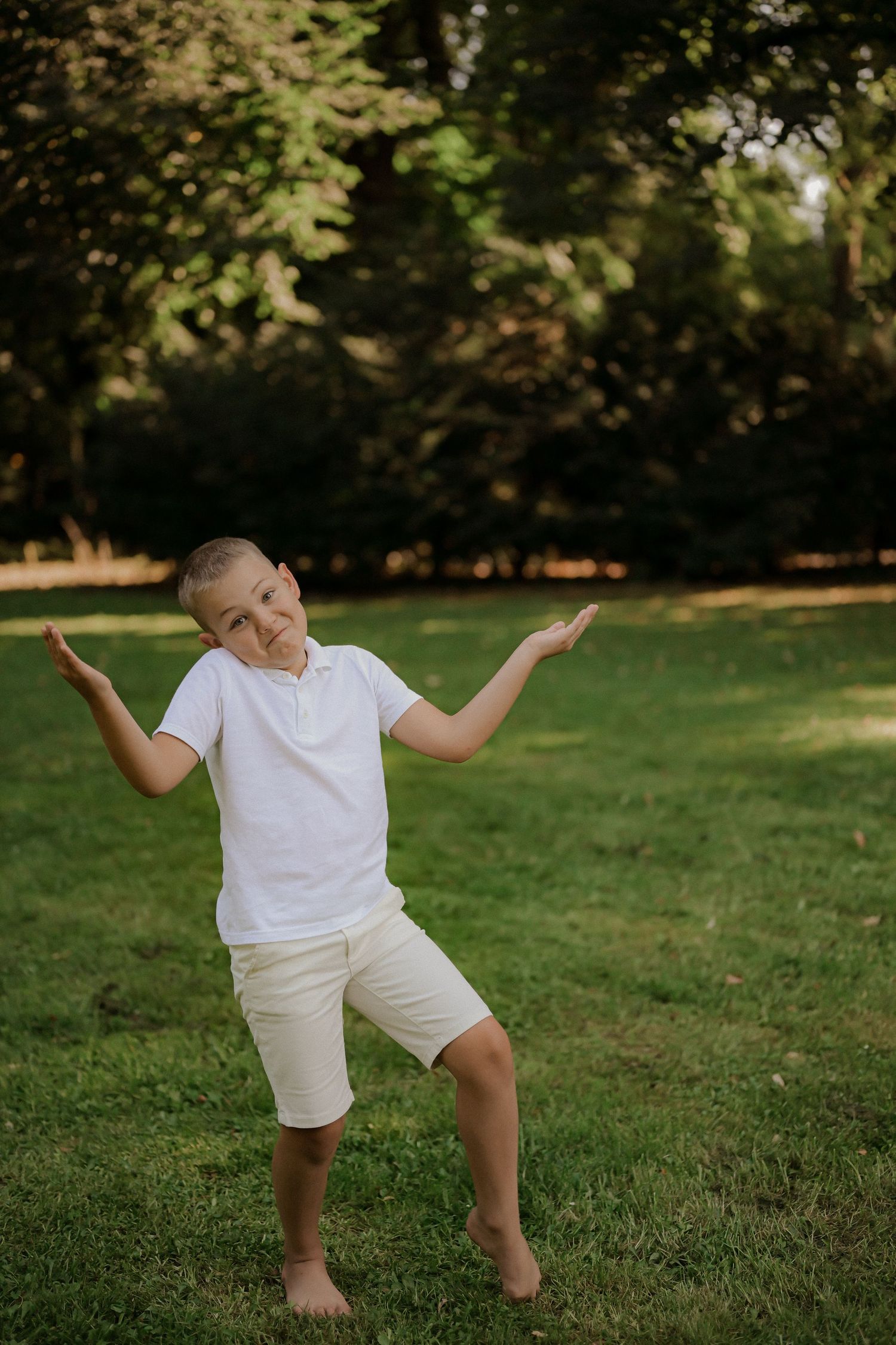 A young boy in white t-shirt and shorts stands barefoot on grass in a park, gesturing with both hands raised.