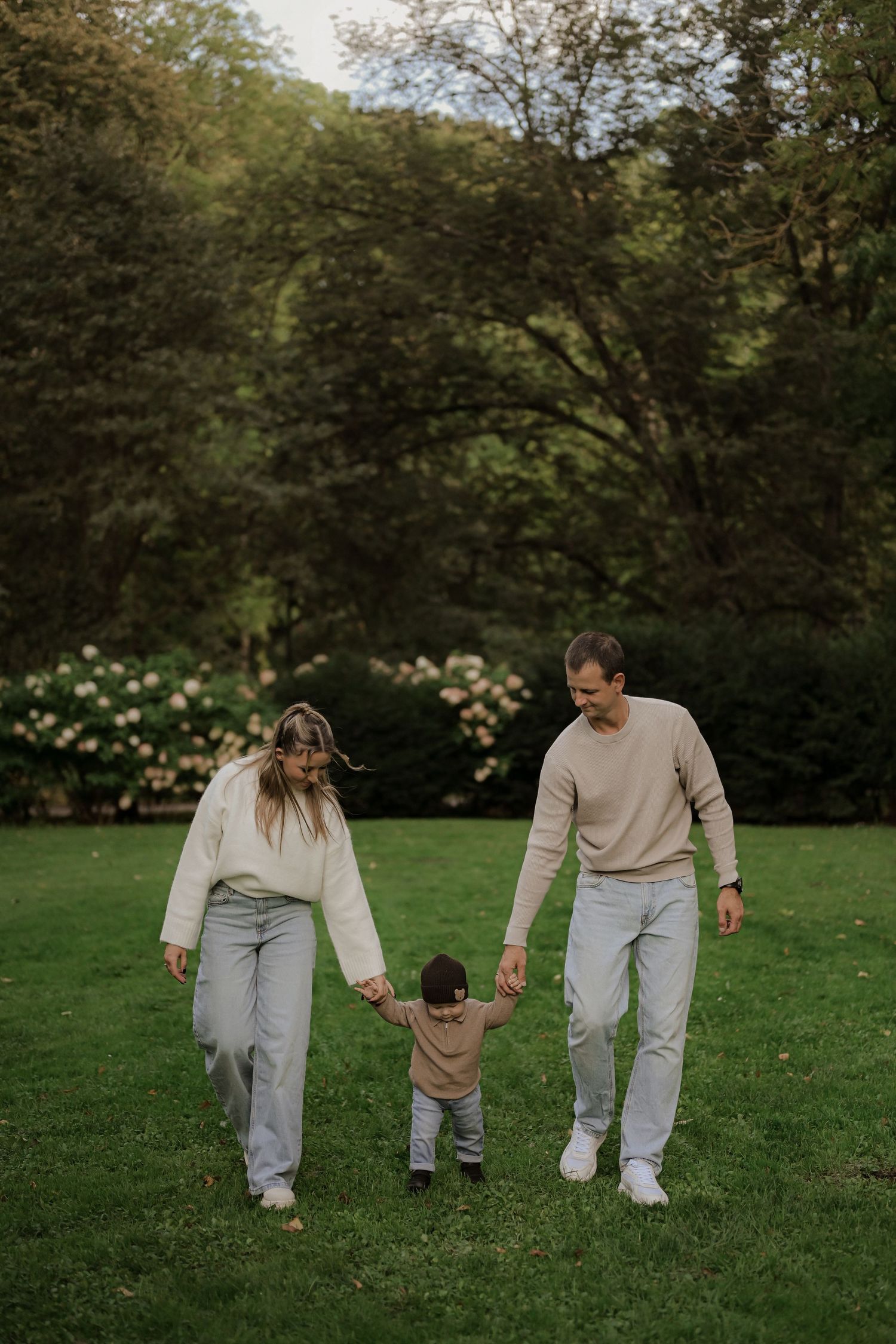 A young family holding hands with their toddler while walking on a green lawn in a lush park.