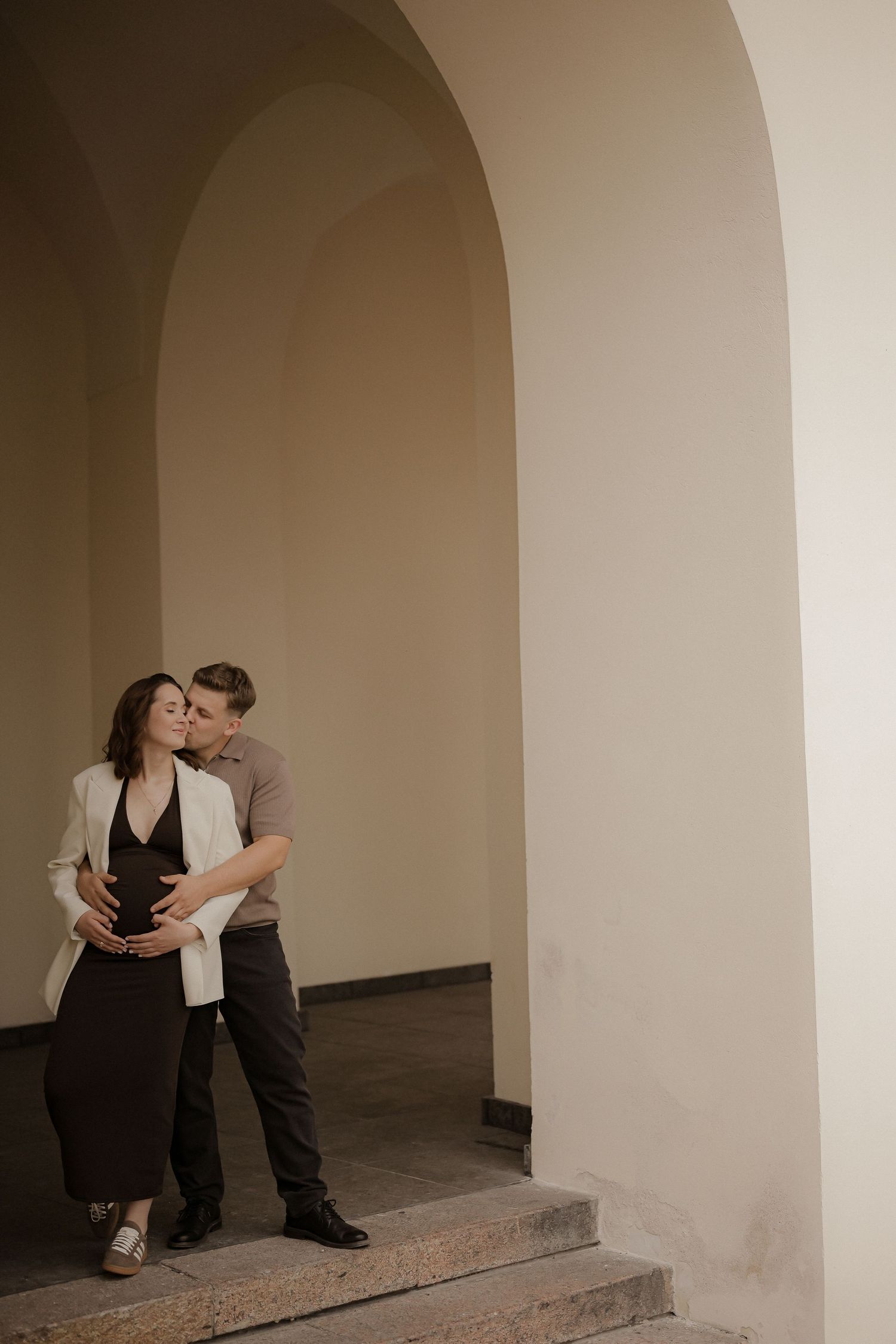A man tenderly embraces a pregnant woman under elegant arched columns.