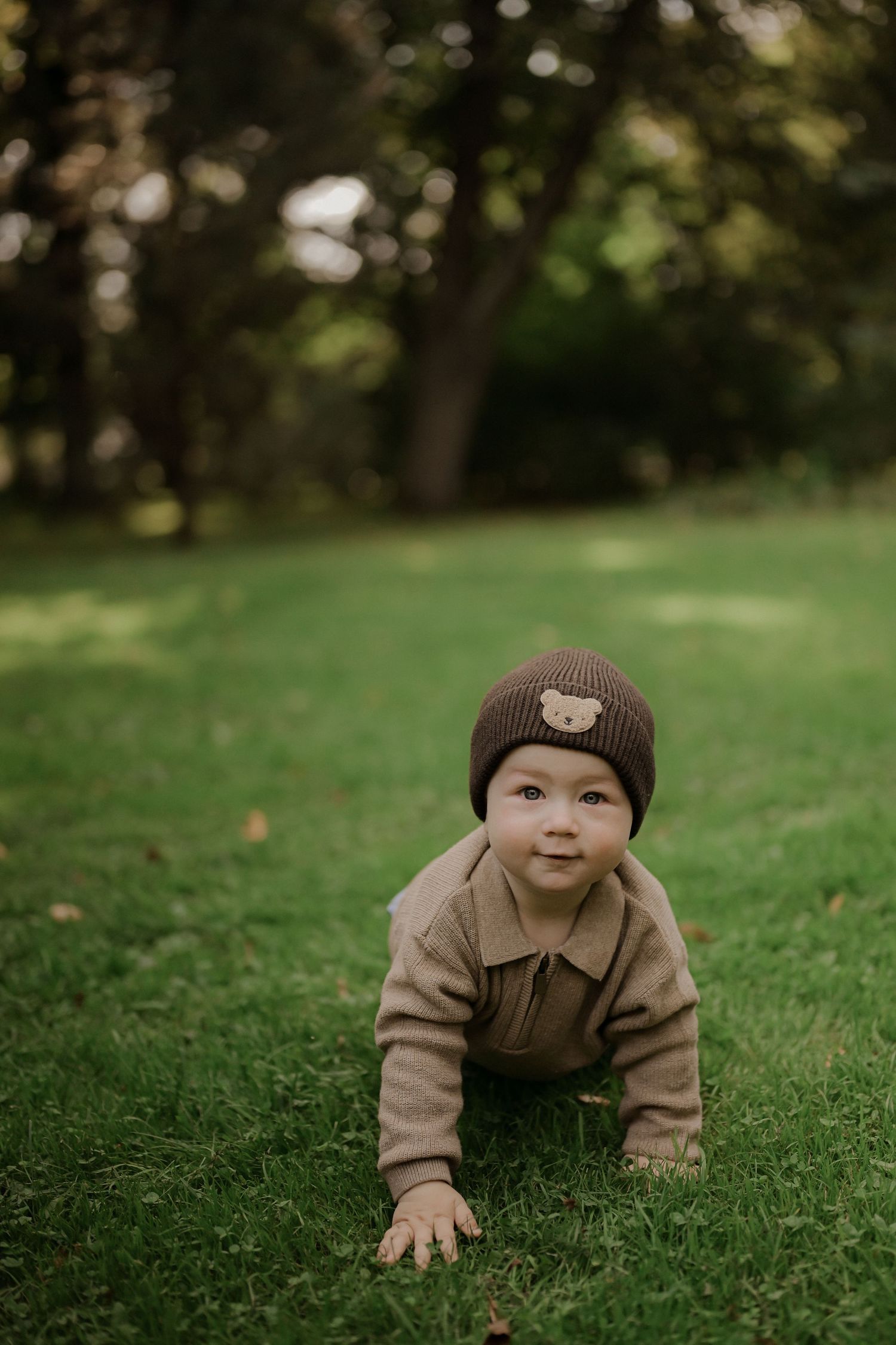 Adorable baby in brown bear beanie and outfit crawling on lush green grass in a park setting.